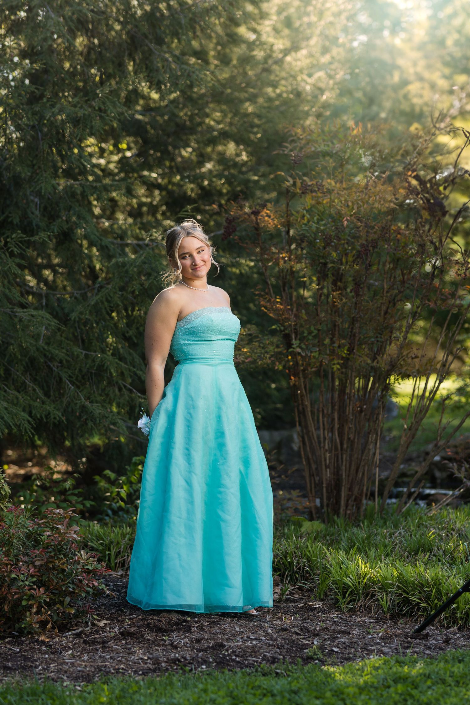 Young woman in a strapless teal prom gown poses gracefully in a lush garden with sunlit trees.