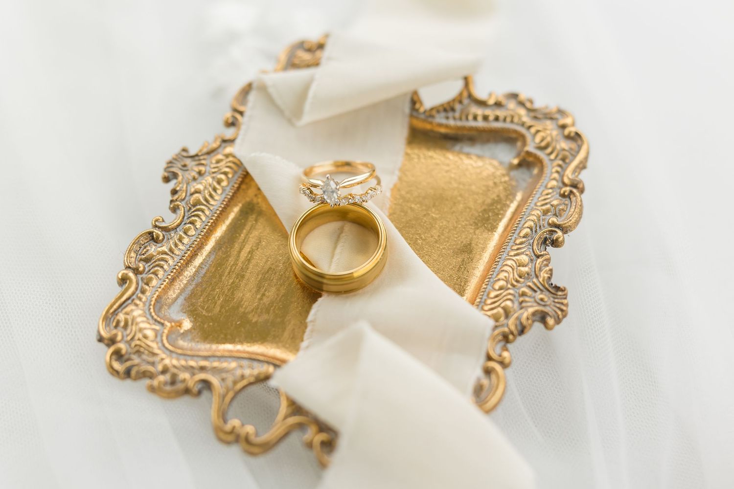 Gold wedding bands and diamond engagement ring displayed on ornate golden trays with white ribbon.