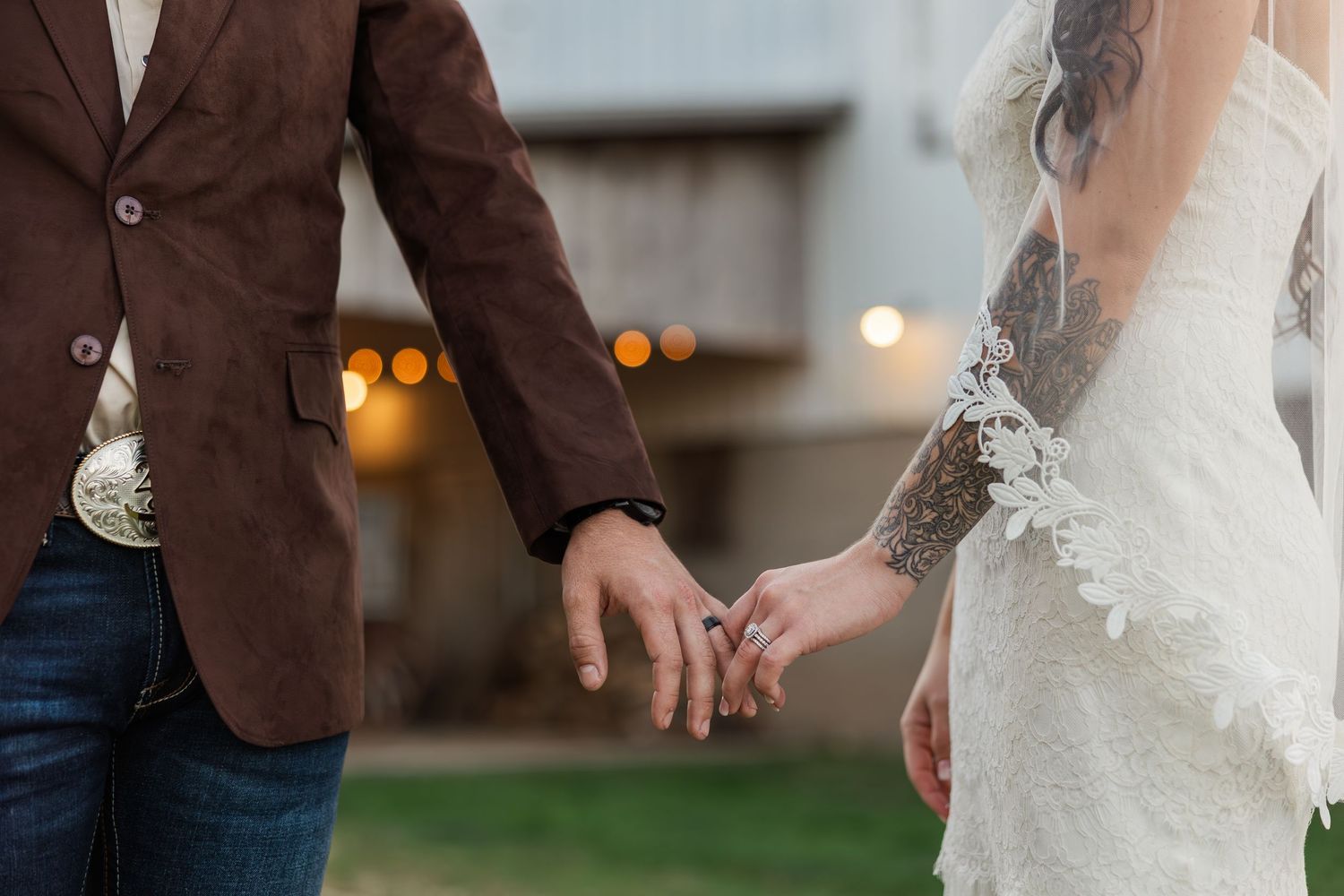 Bride and groom holding hands, wearing lace wedding dress and brown blazer with jeans at outdoor wedding.