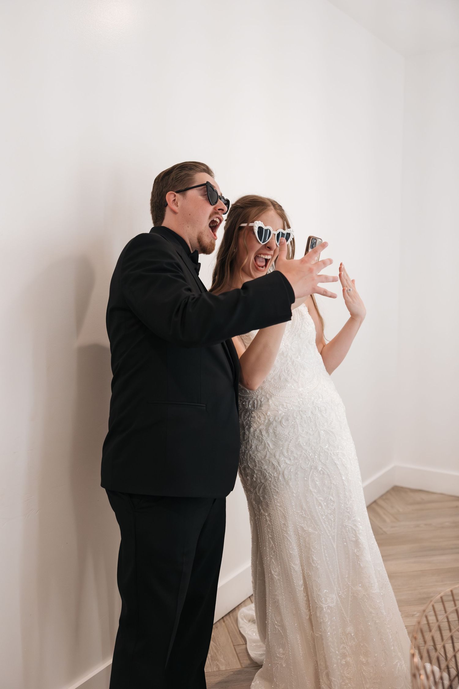 A couple in wedding attire wearing sunglasses pose playfully against a white wall.