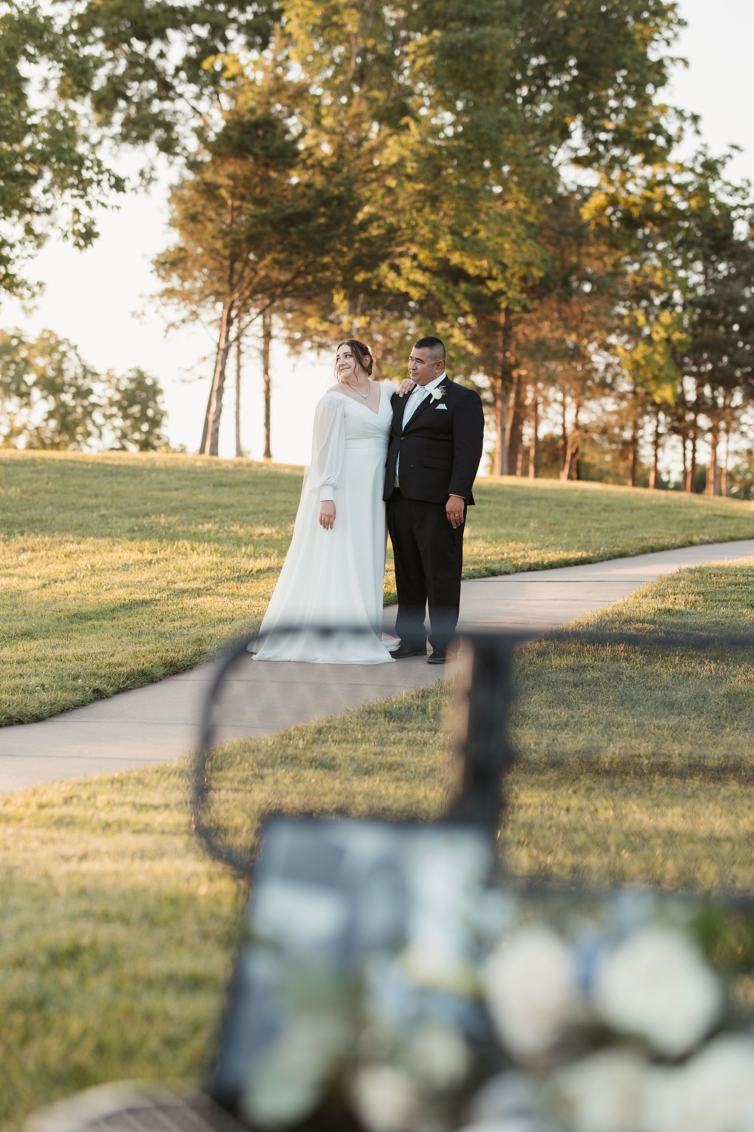 Couple poses on pathway while photographer captures moment through bench frame.