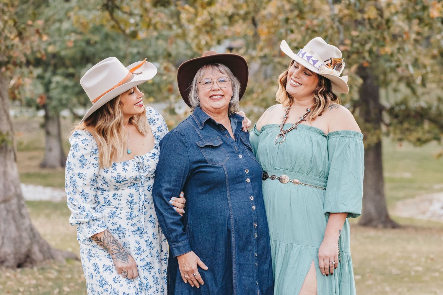 Three women in cowboy hats and dresses pose together outdoors on a sunny autumn day.
