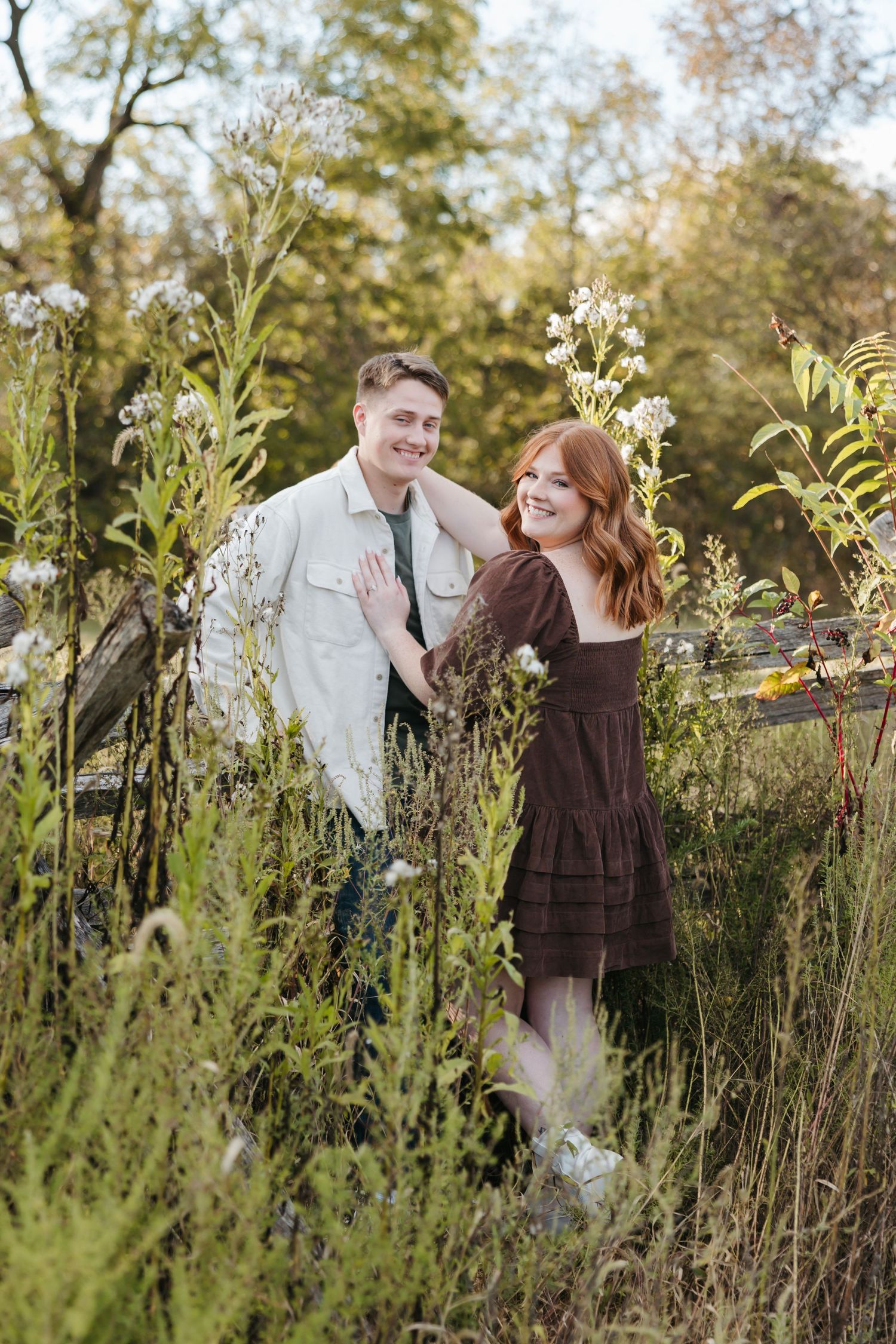 Couple in casual attire pose playfully among tall wildflowers at sunset.