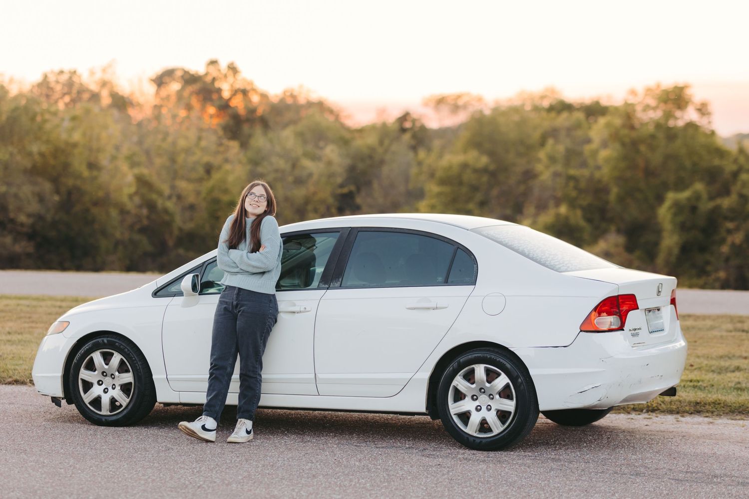 Person standing beside white sedan car in an open field during sunset.
