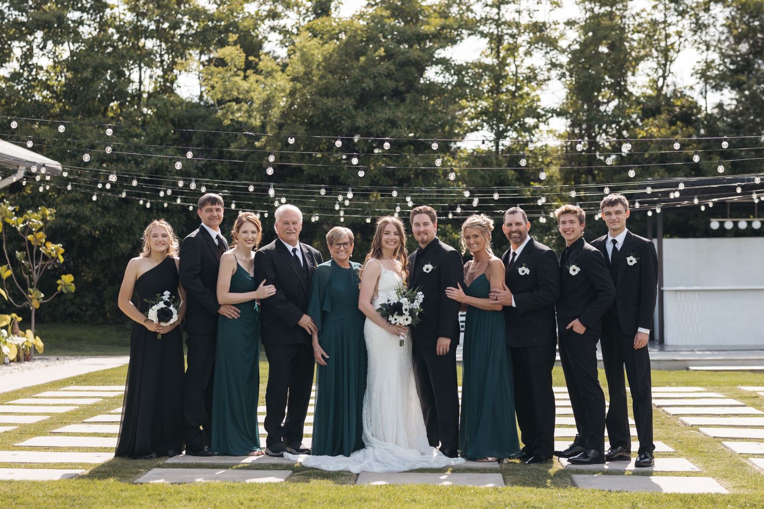 Wedding party in black and white attire poses together on checkerboard lawn under string lights.