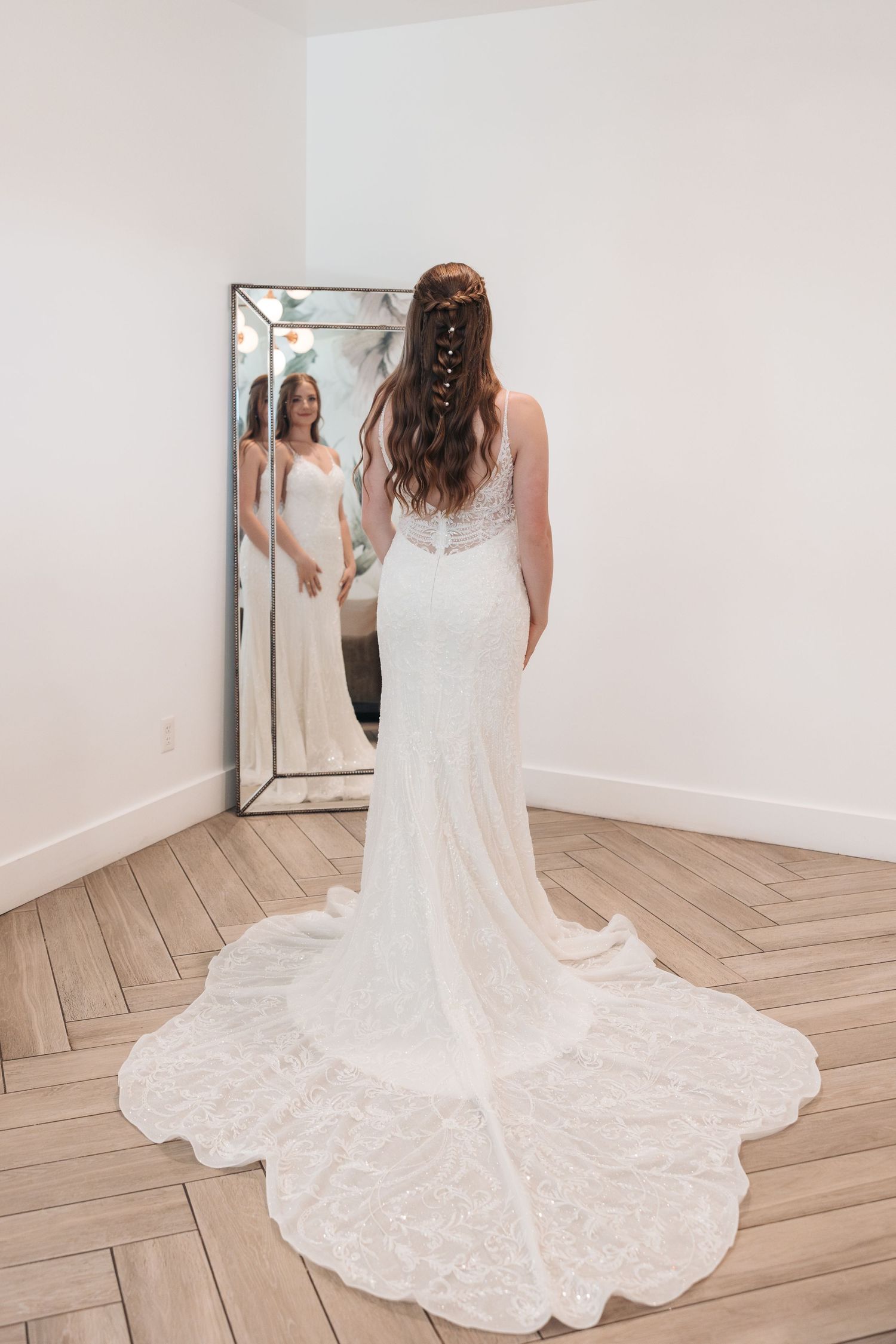 A bride in a flowing lace wedding dress with train stands in front of a decorative mirror on wooden flooring.