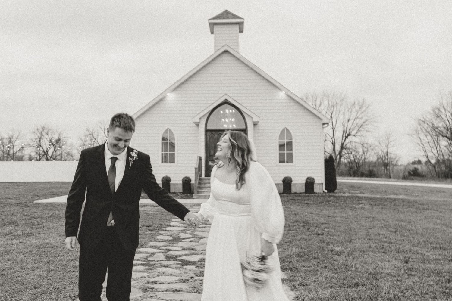 Wedding couple walks hand in hand along a path leading to a white country church.