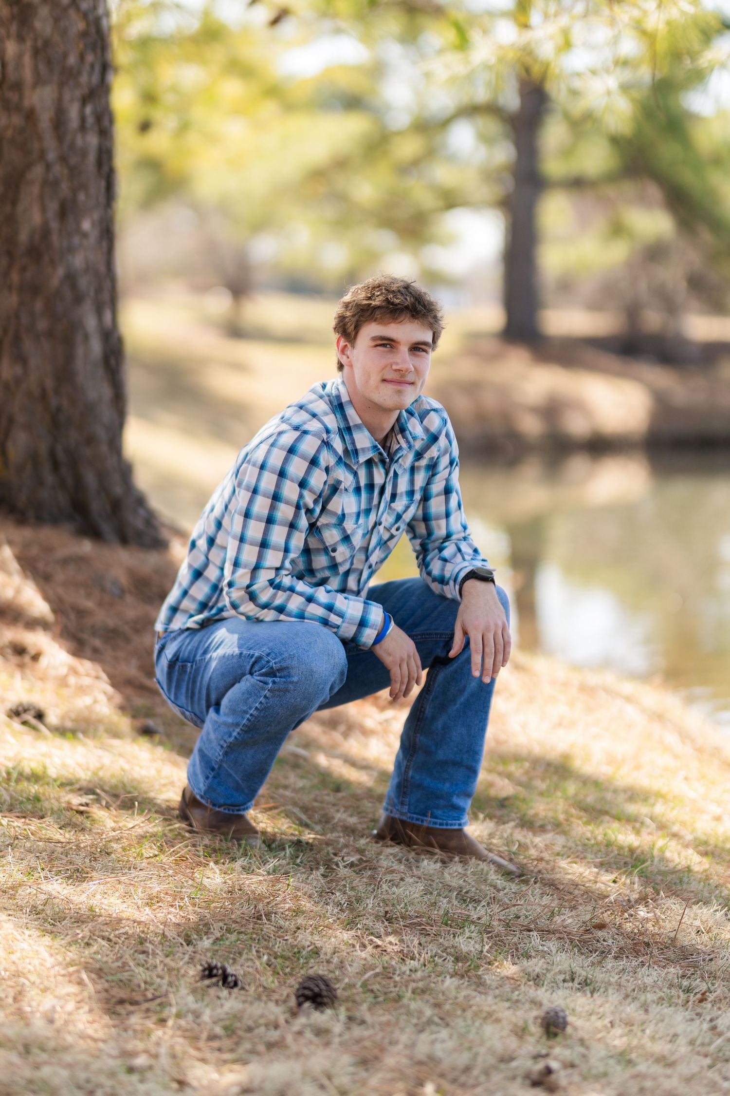 Outdoor portrait by water with person crouching in blue plaid shirt and jeans.