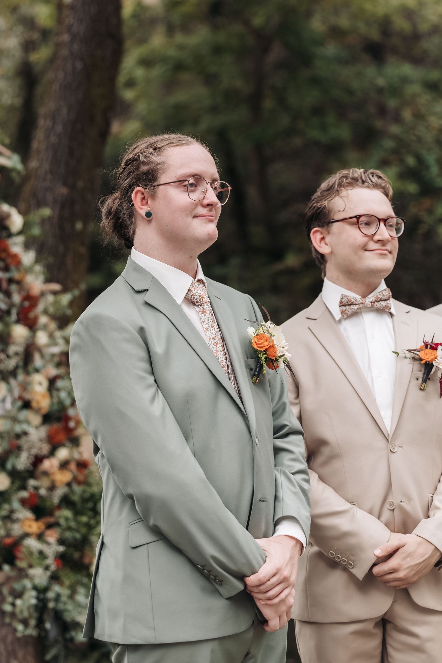 Two wedding party members in coordinating suits and glasses stand together during ceremony.