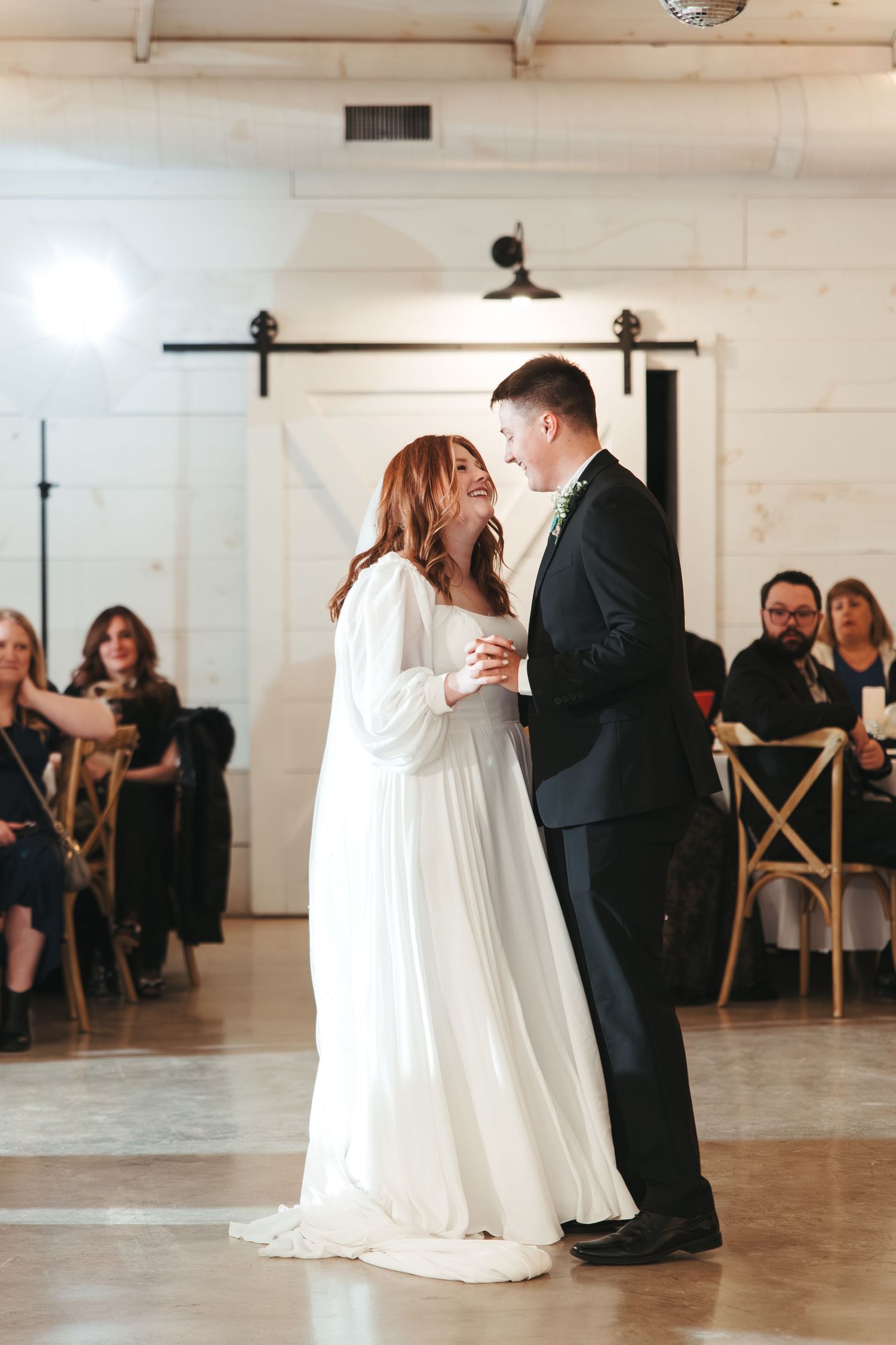 A couple dances together in a modern white venue with wooden chairs and guests watching.