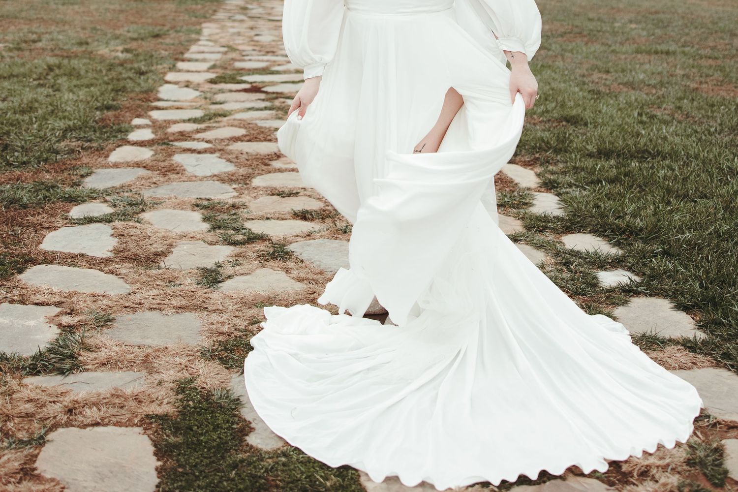 Flowing white wedding dress train on stone pathway through grassy field.