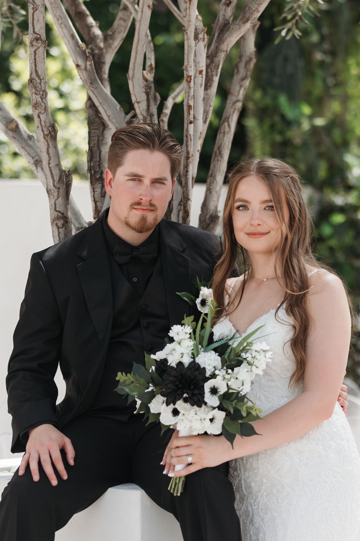 Wedding portrait with bride in white lace gown and groom in black suit holding white floral bouquet.