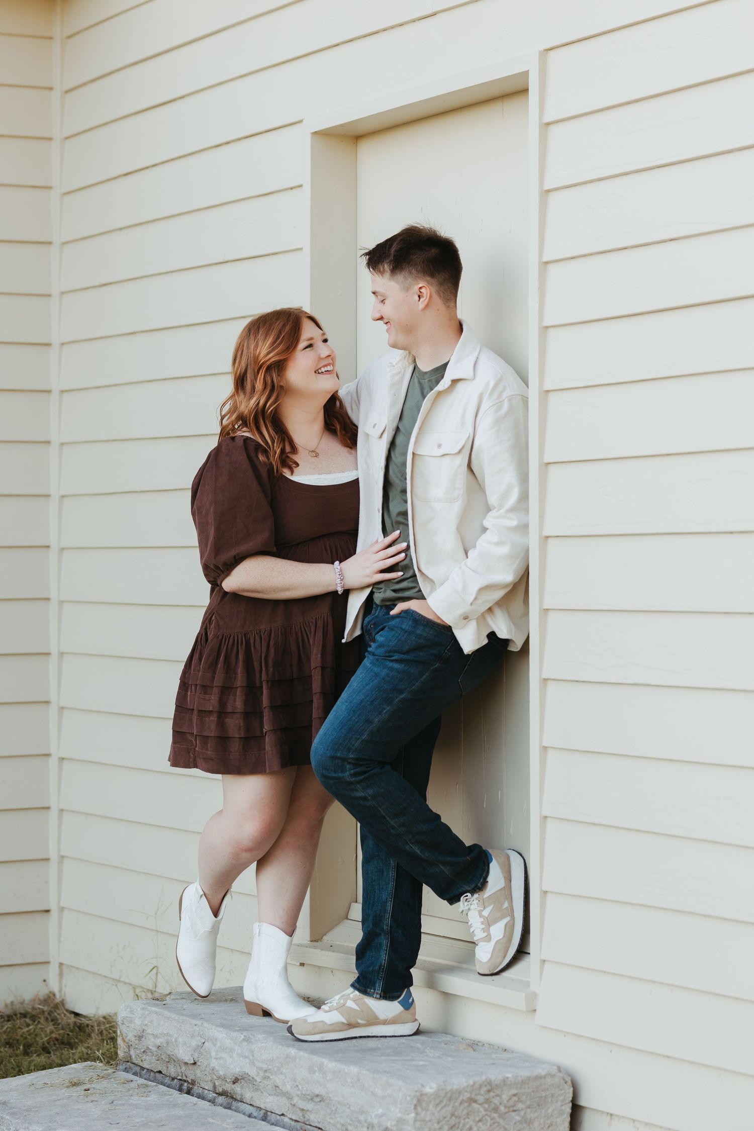 Couple in casual outfits pose playfully against white house exterior wall.