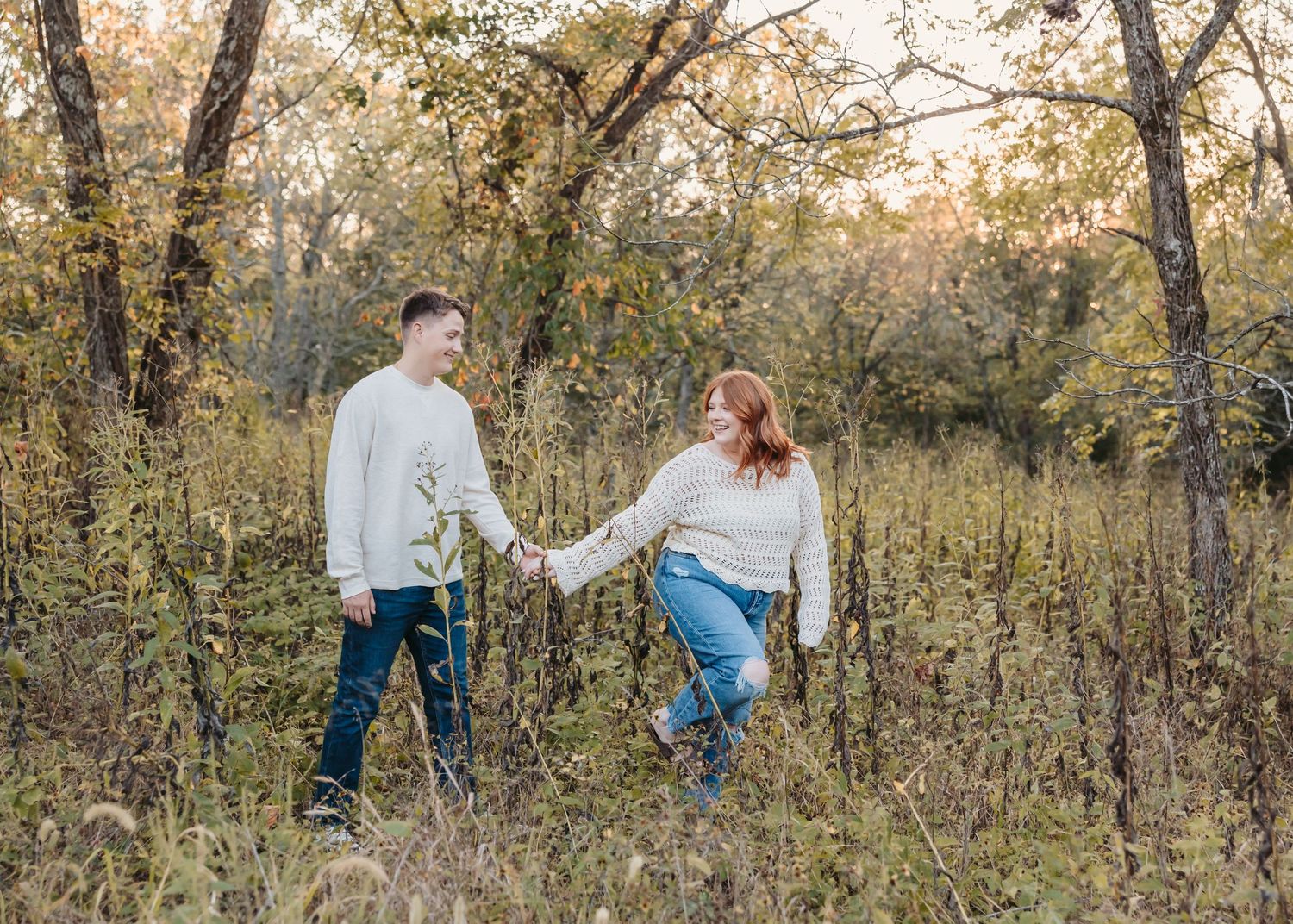 Two people hold hands while walking through a field of tall grass at sunset.