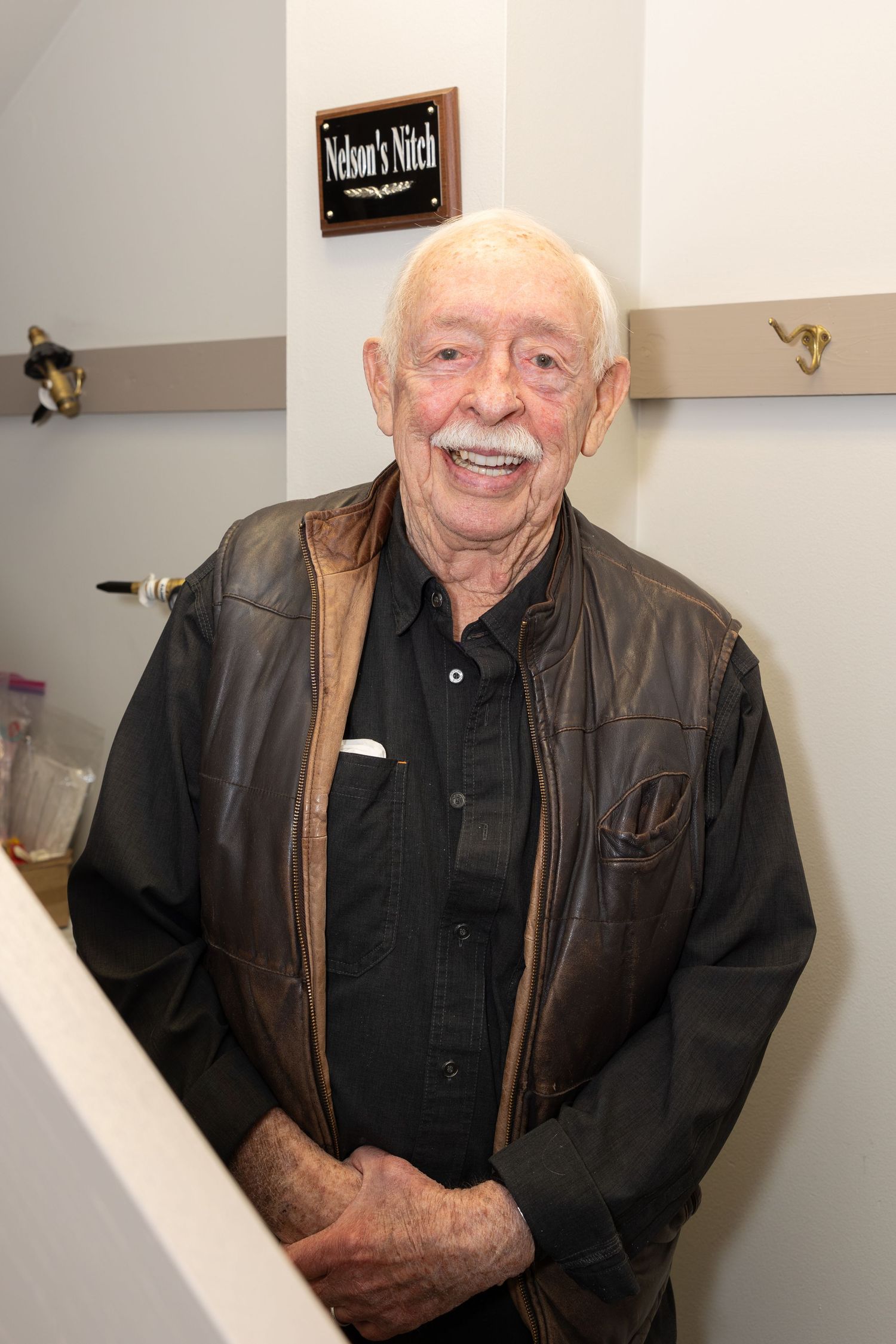 Senior guest in leather jacket stands smiling in hallway.