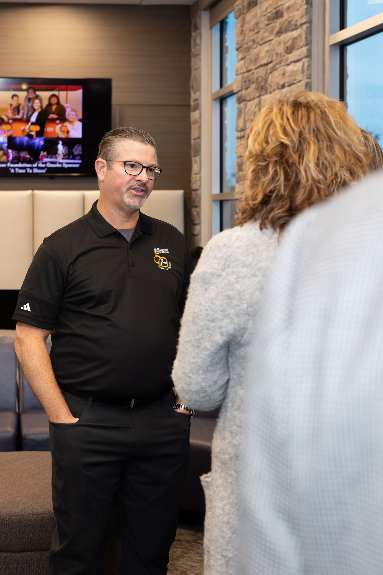 Person in black polo shirt with logo speaking to someone in office setting.
