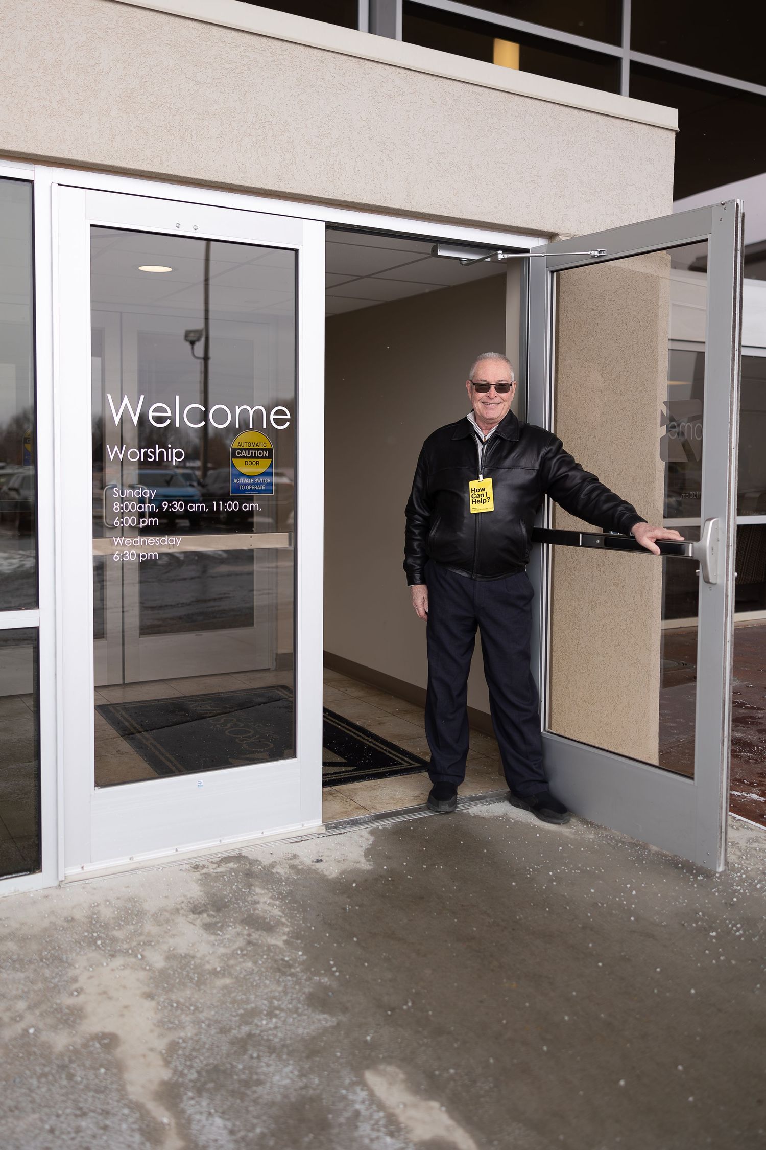 Person holding open glass entrance door with welcome signage at a church building.