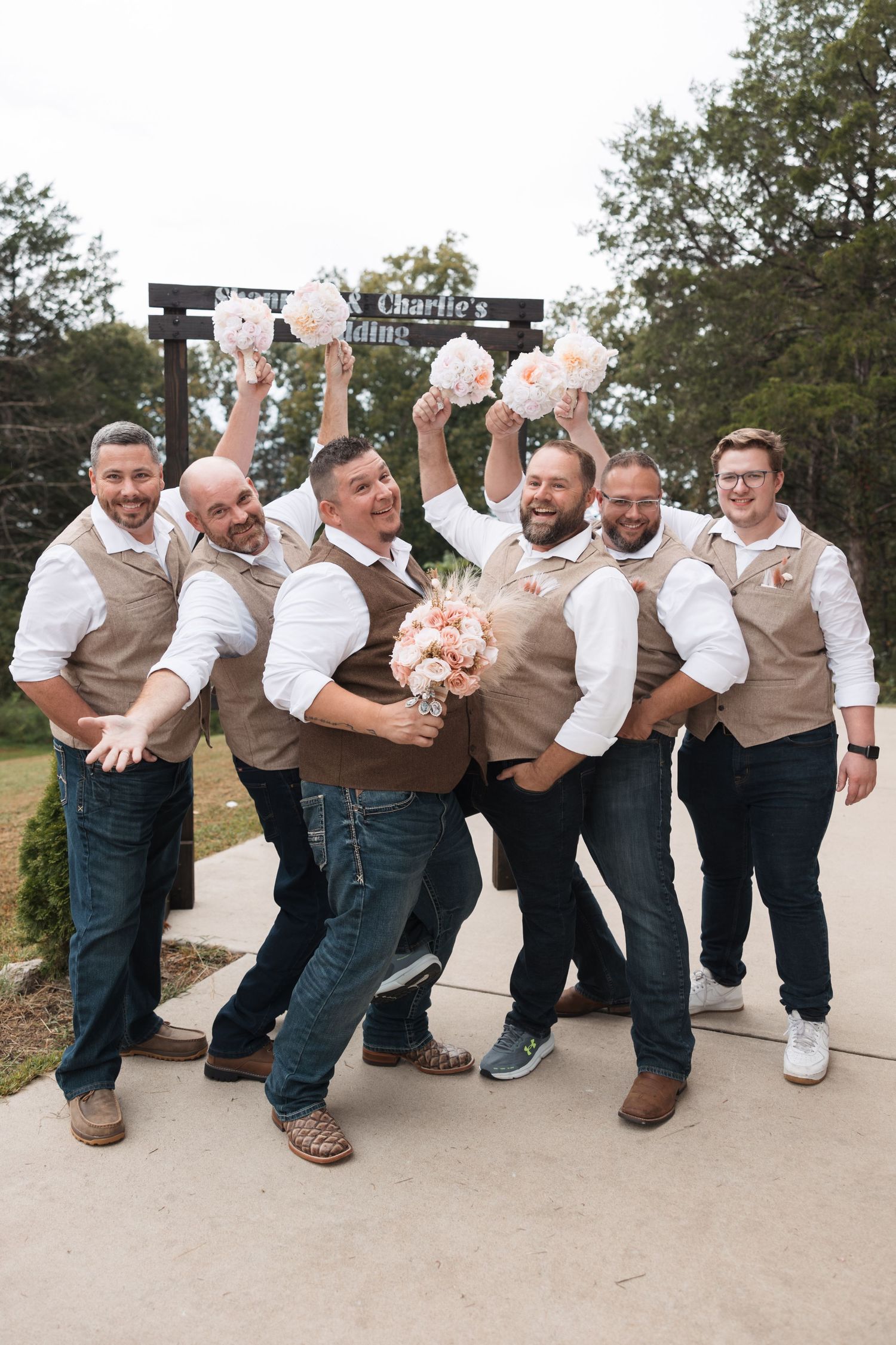 Groomsmen in coordinated vests playfully pose with bouquets on an outdoor pathway.