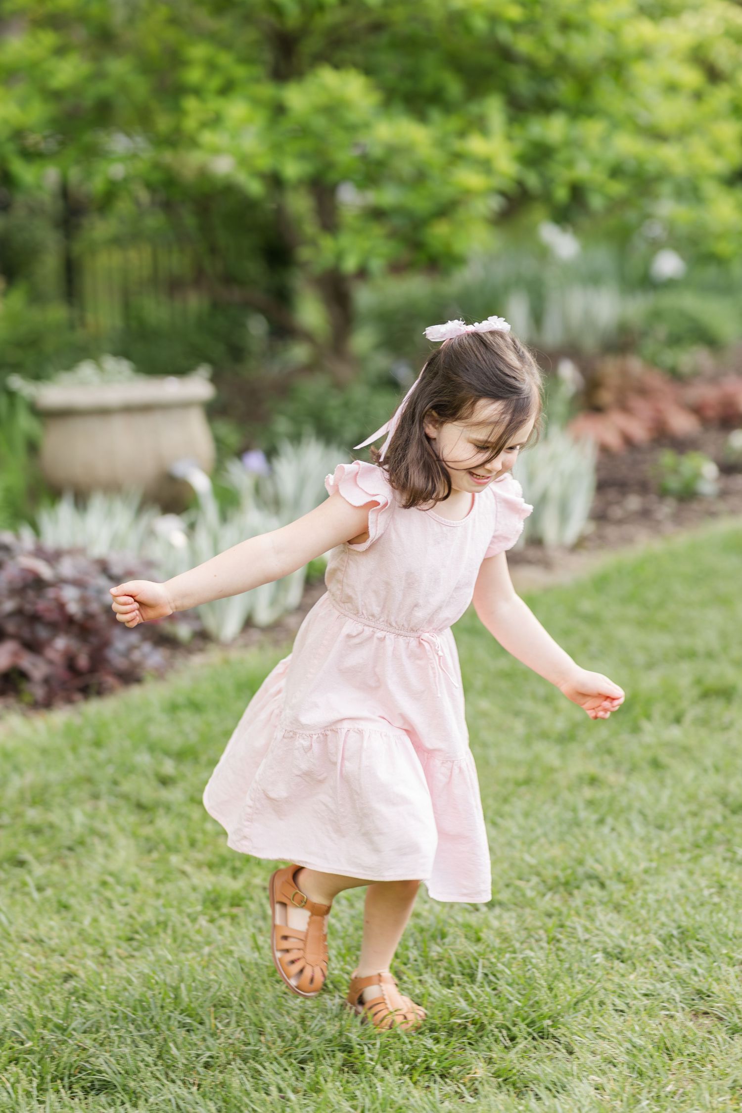 A child in a pink dress twirls and dances across a lush green lawn in a garden setting.