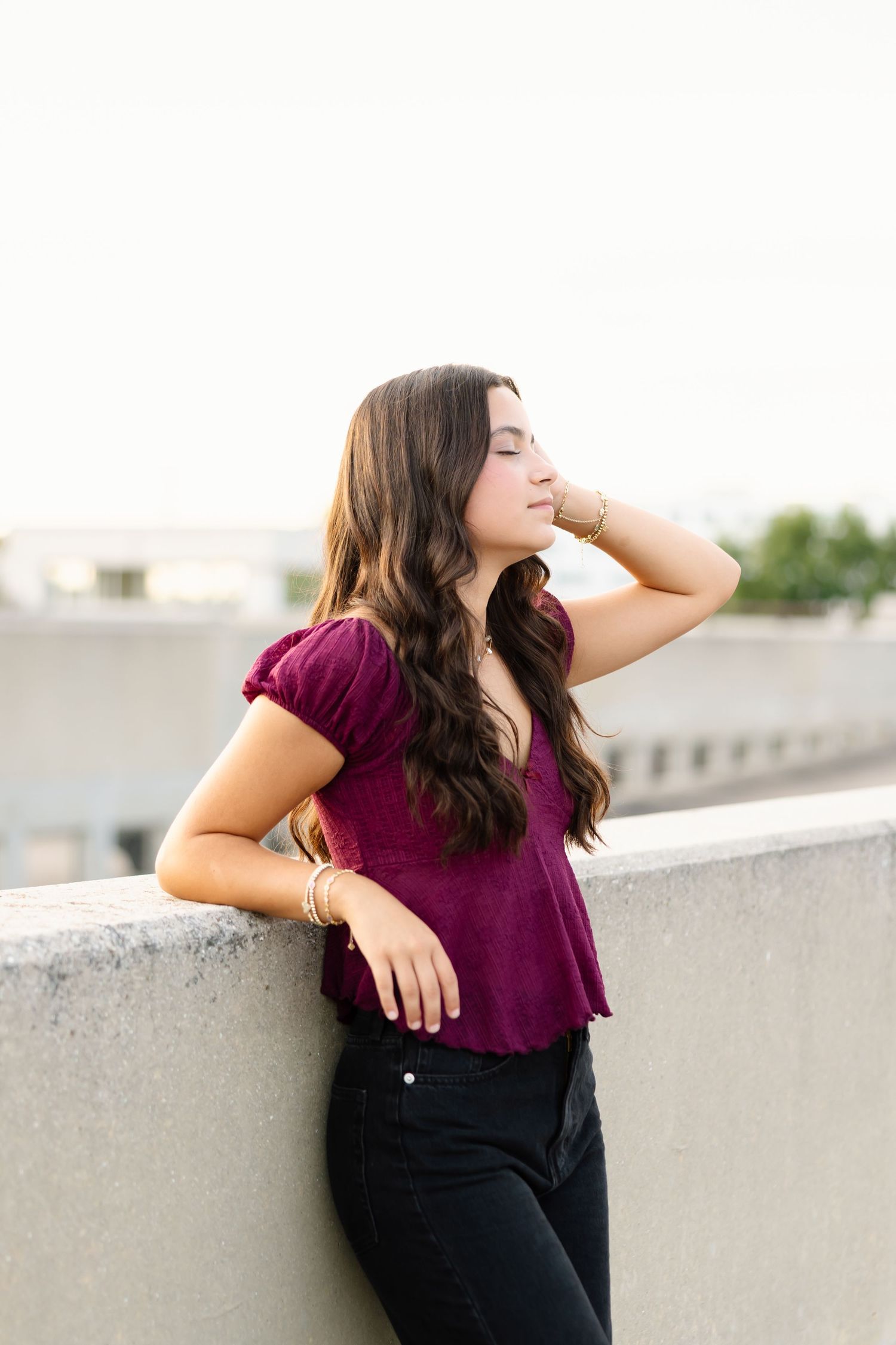A relaxed outdoor portrait against a white wall wearing a burgundy blouse and dark pants.