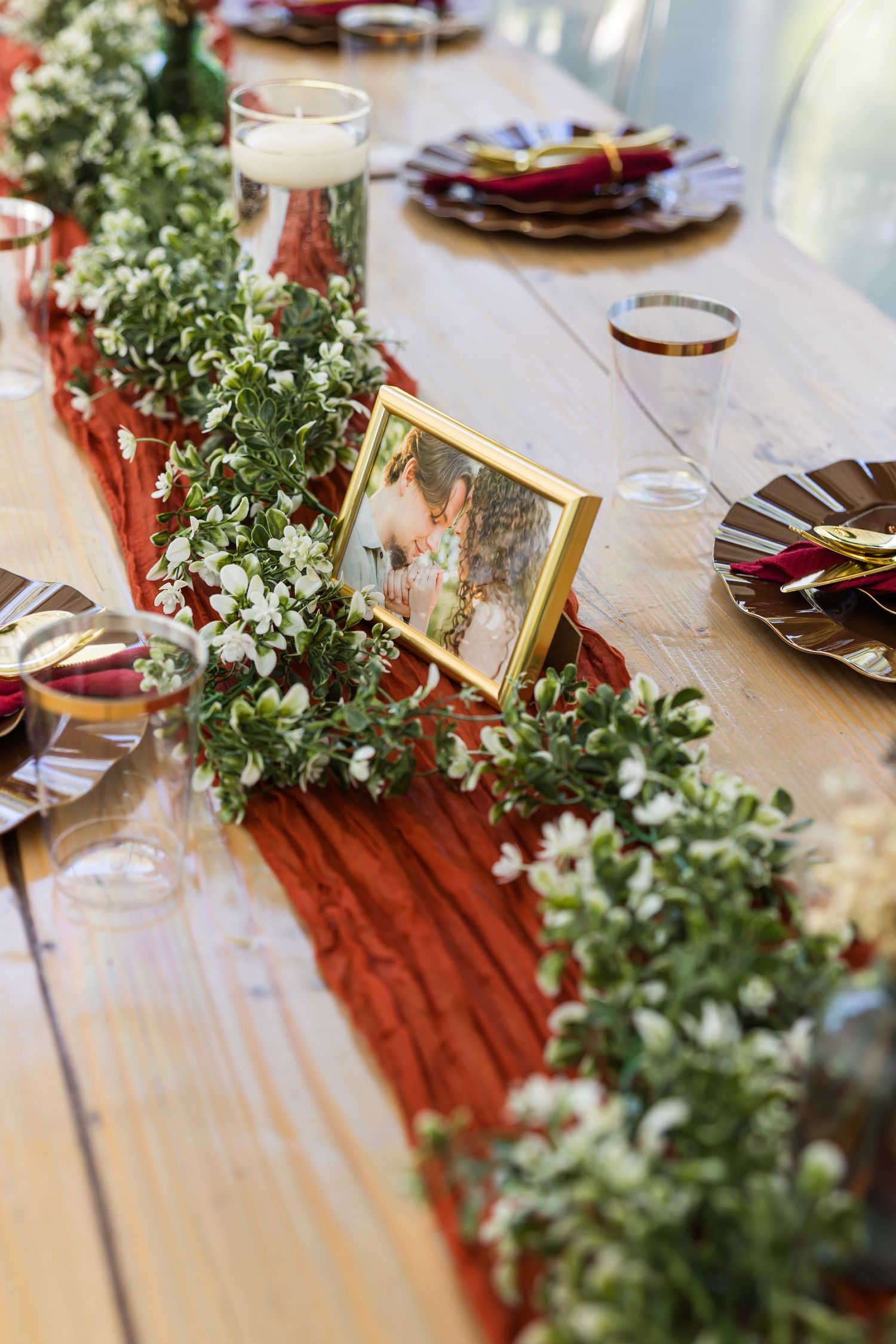 Rustic wedding table with red runner, greenery garland, white flowers, gold-framed photo, and elegant place settings.