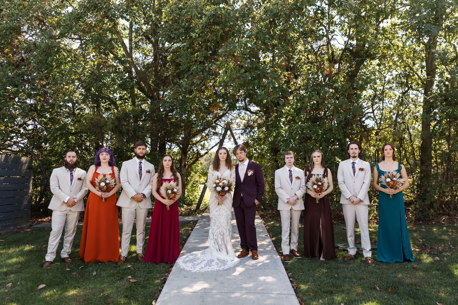 Wedding party in colorful dresses and tan suits stands together outdoors before tree-lined aisle.