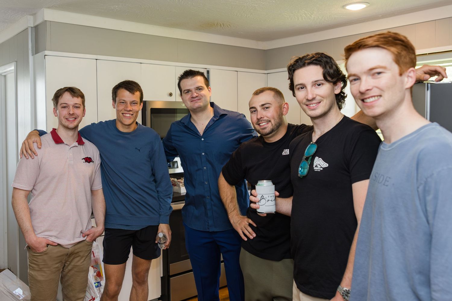 Group of six smiling young men posing together in a kitchen during a casual gathering.