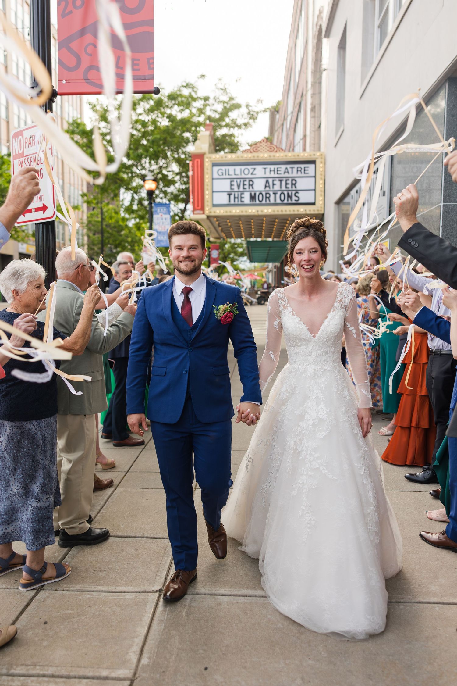 Newlyweds Mr. and Mrs. Morton walk hand-in-hand through a ribbon wand sendoff outside a theater.