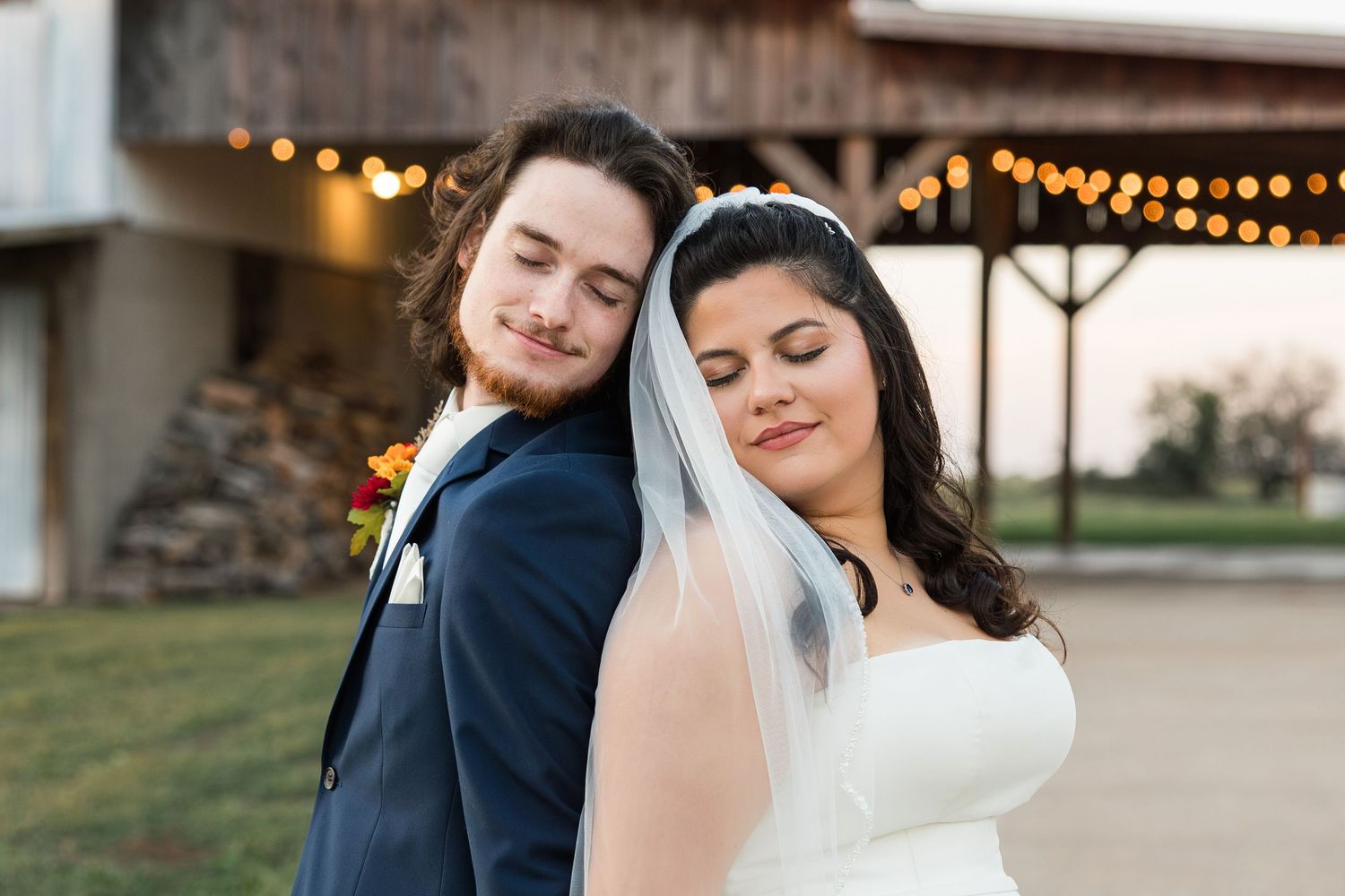 Bride and groom share a tender back-to-back pose outdoors at a rustic barn wedding venue with warm string lights.