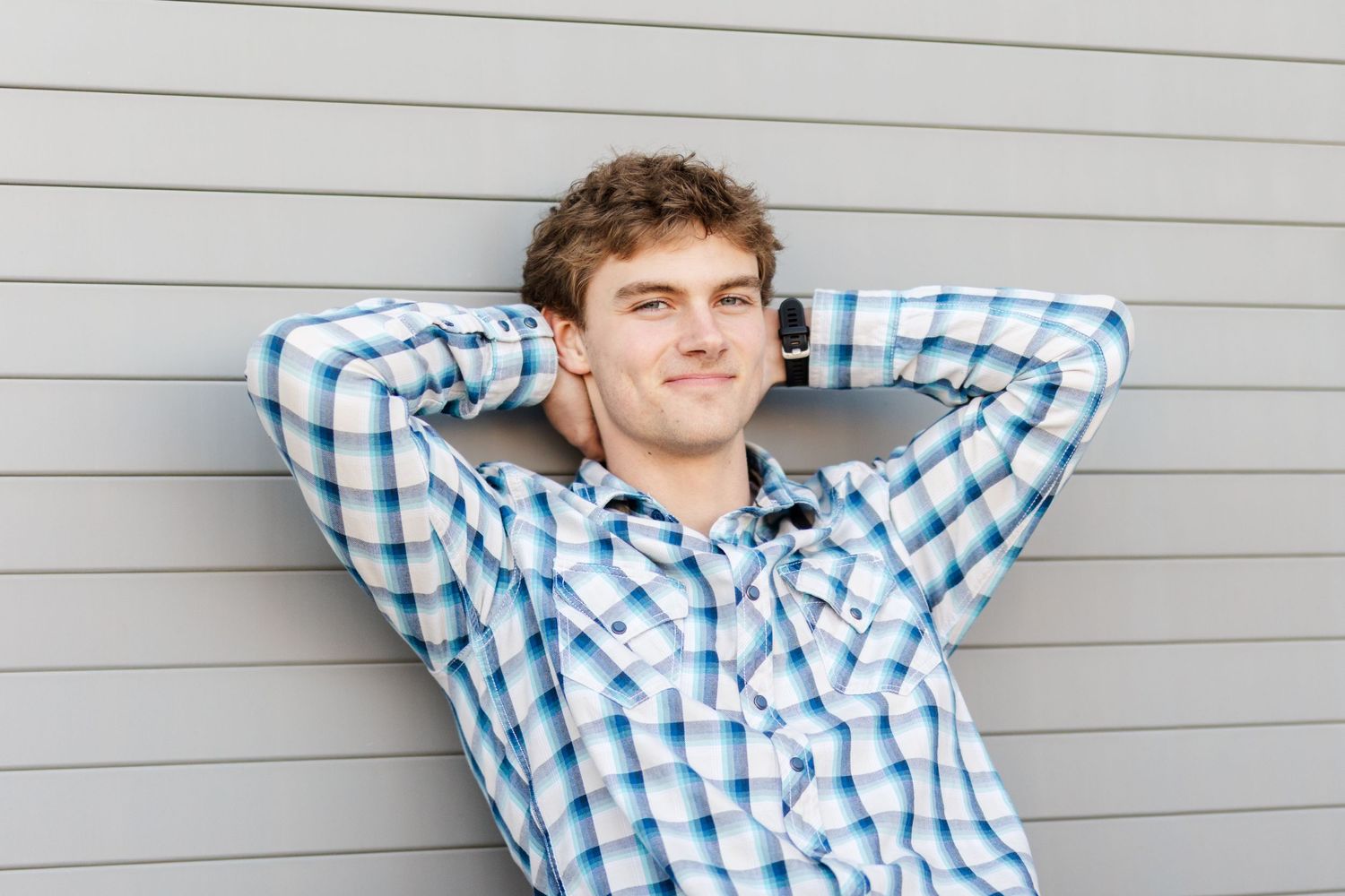 A person wearing a blue checkered shirt leans back relaxed against a gray wall with hands behind their head.