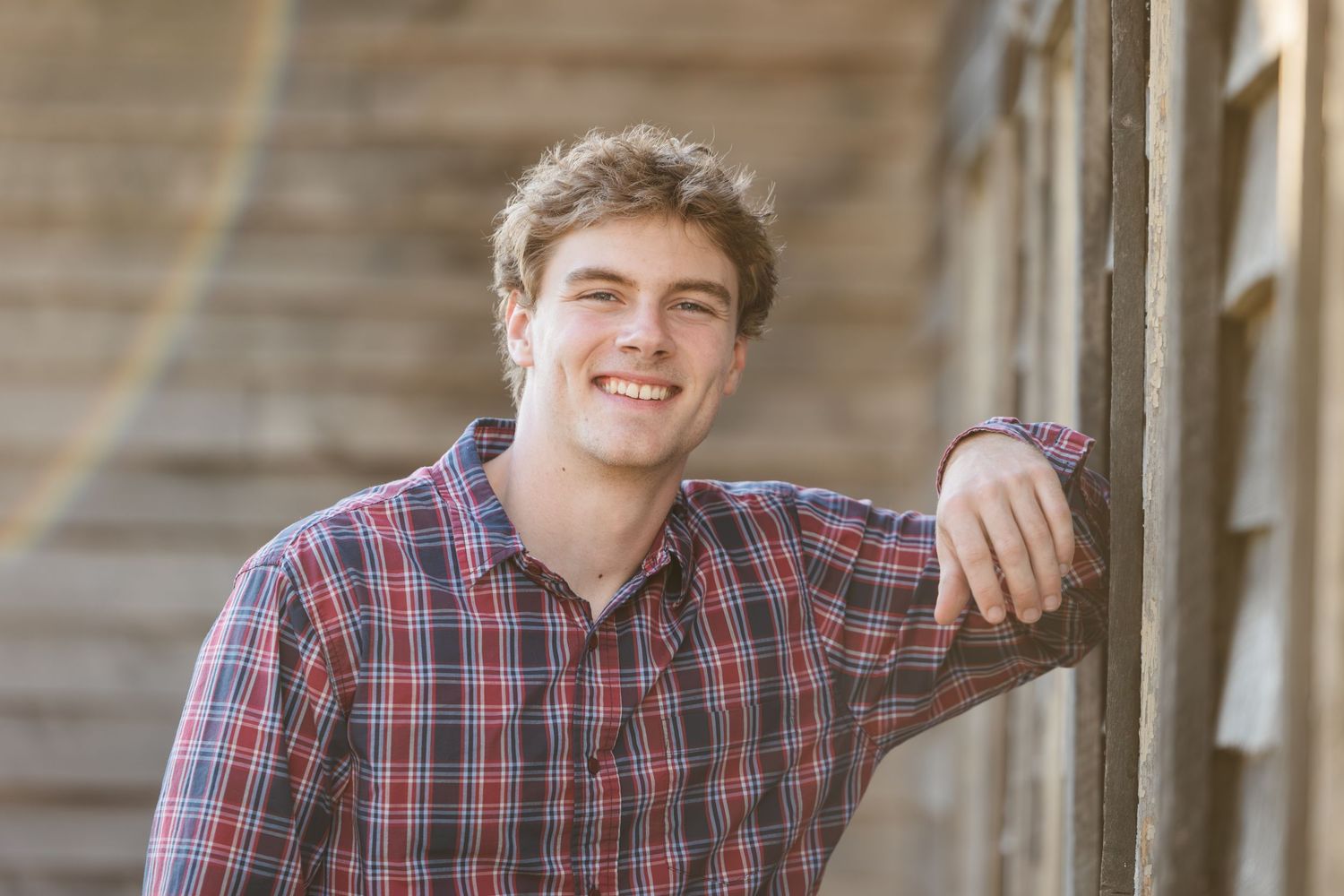 A young person in a red plaid button-down shirt leans casually against a wooden wall with natural lighting.