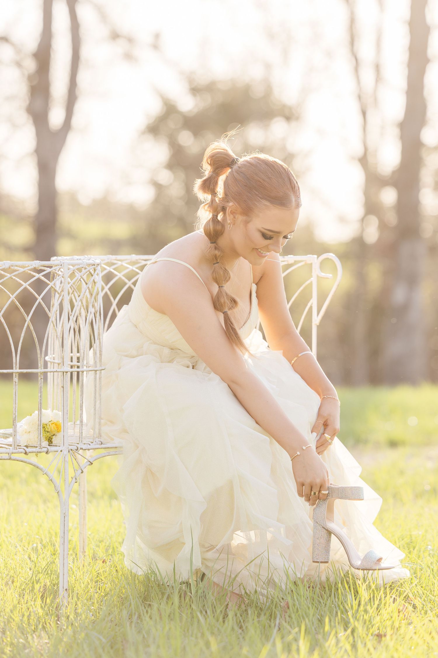 A bride in a white wedding dress adjusts her boots while sitting on a vintage white metal bench in golden sunlight.