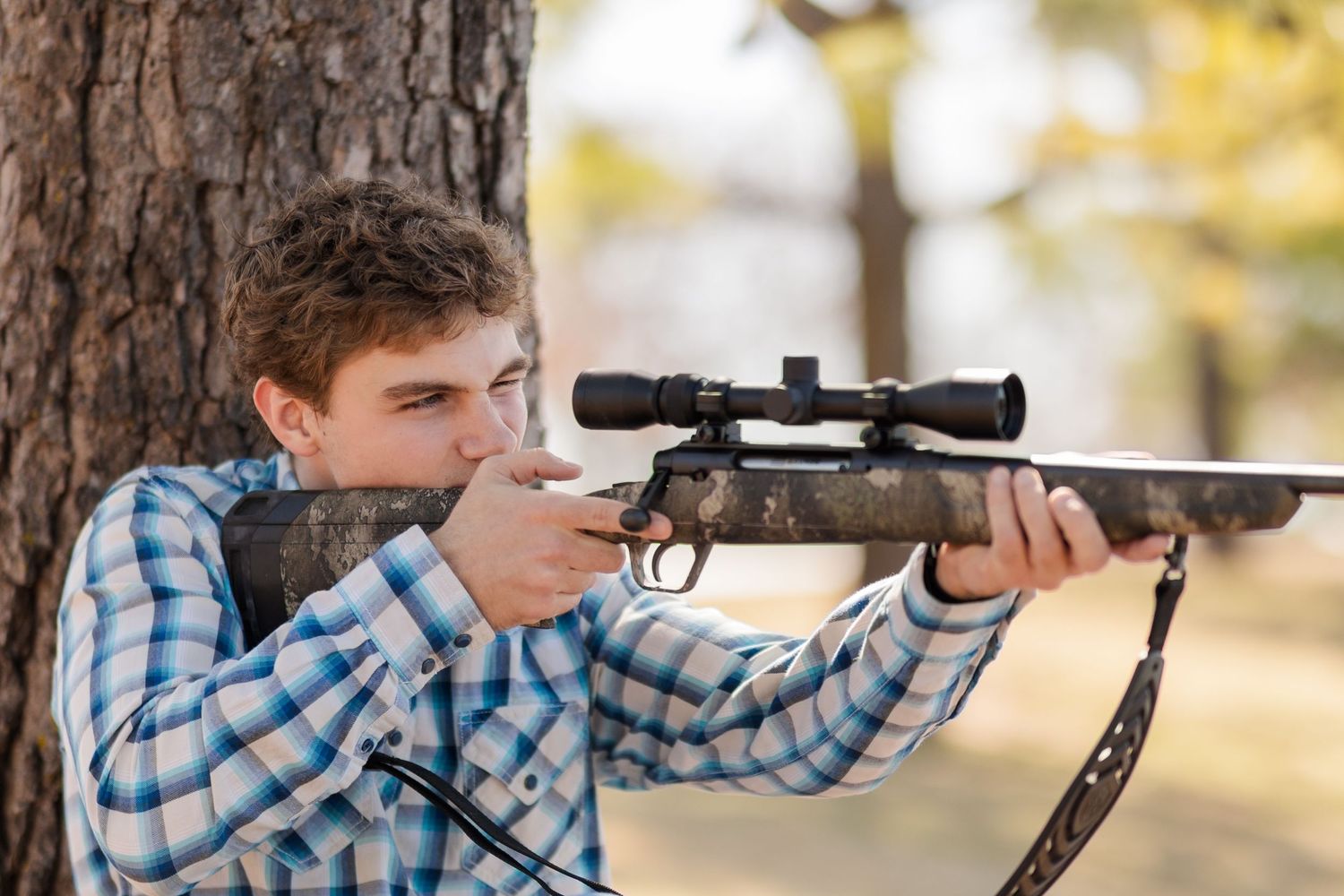 A person in a plaid shirt takes aim with a scoped hunting rifle while leaning against a tree trunk.