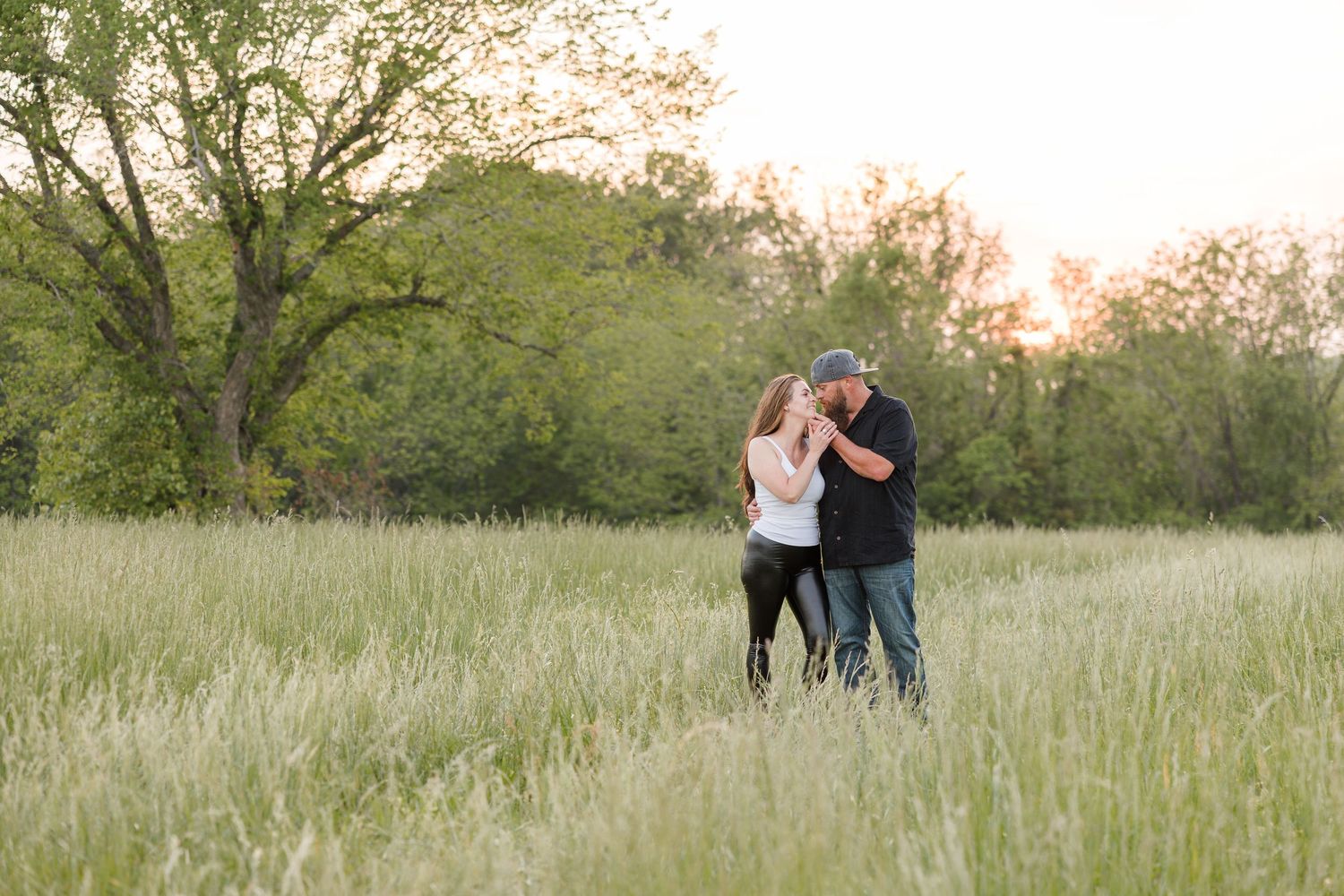 A couple embraces while standing in a sunlit meadow with tall grass and trees during golden hour.
