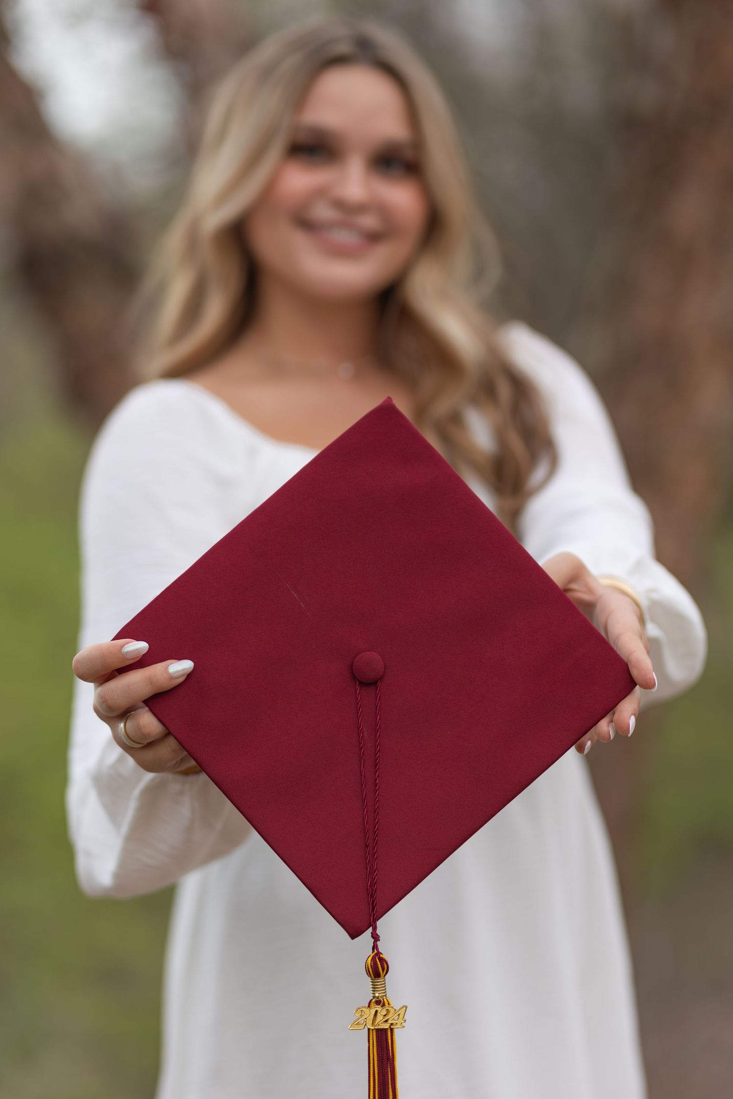 A maroon graduation cap with gold tassel is held up against an outdoor background with soft bokeh.