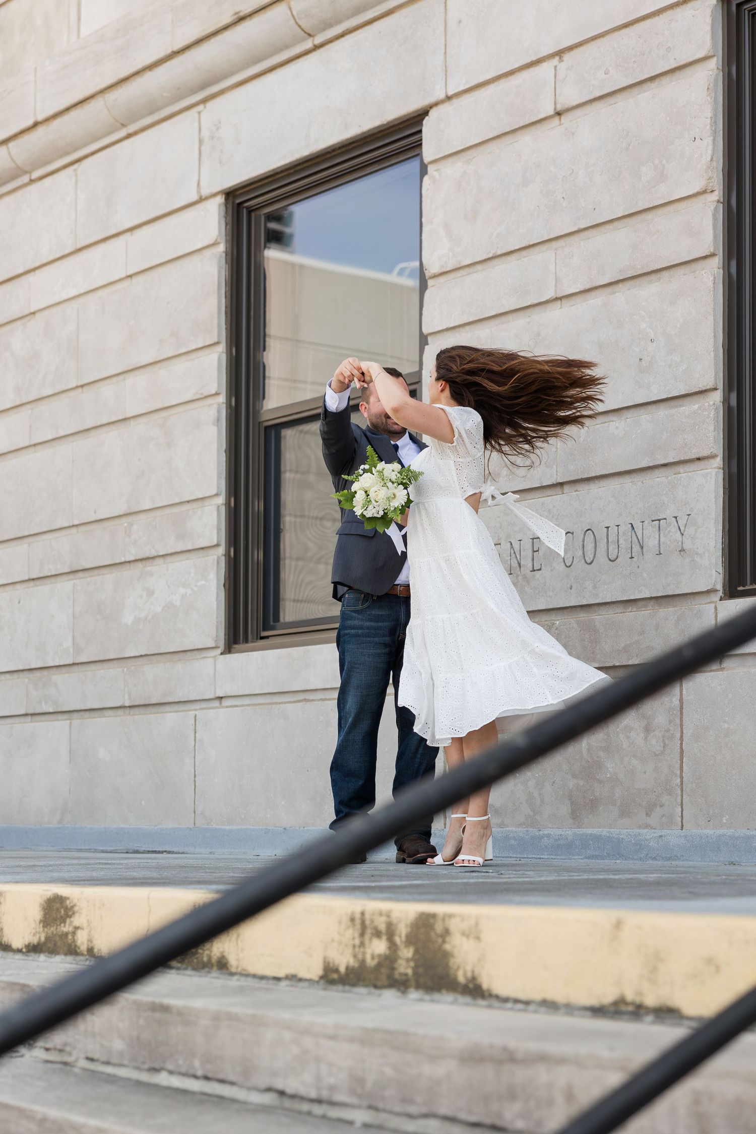 A couple in wedding attire share a romantic dance moment on stone steps outside a county building.