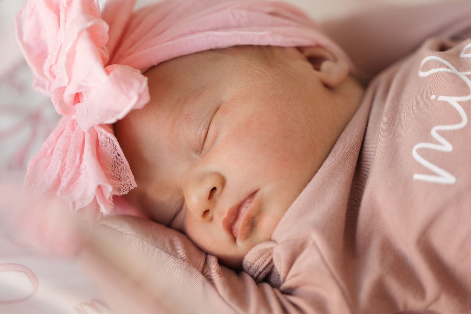 Peaceful sleeping baby wrapped in soft pink blankets with tulle bow headband lying on plush bedding.