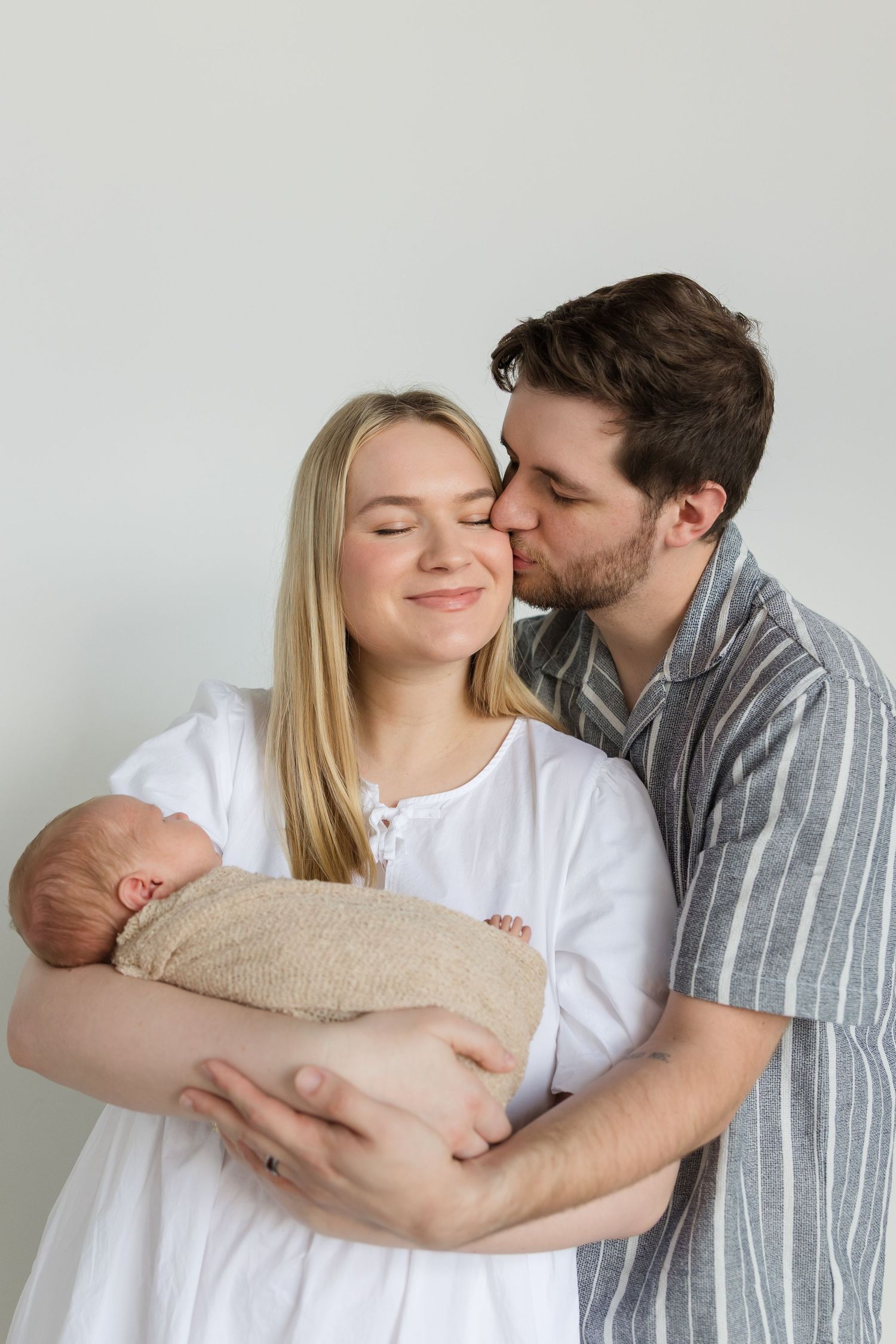 A couple shares intimate moments while holding their newborn baby against a white background.