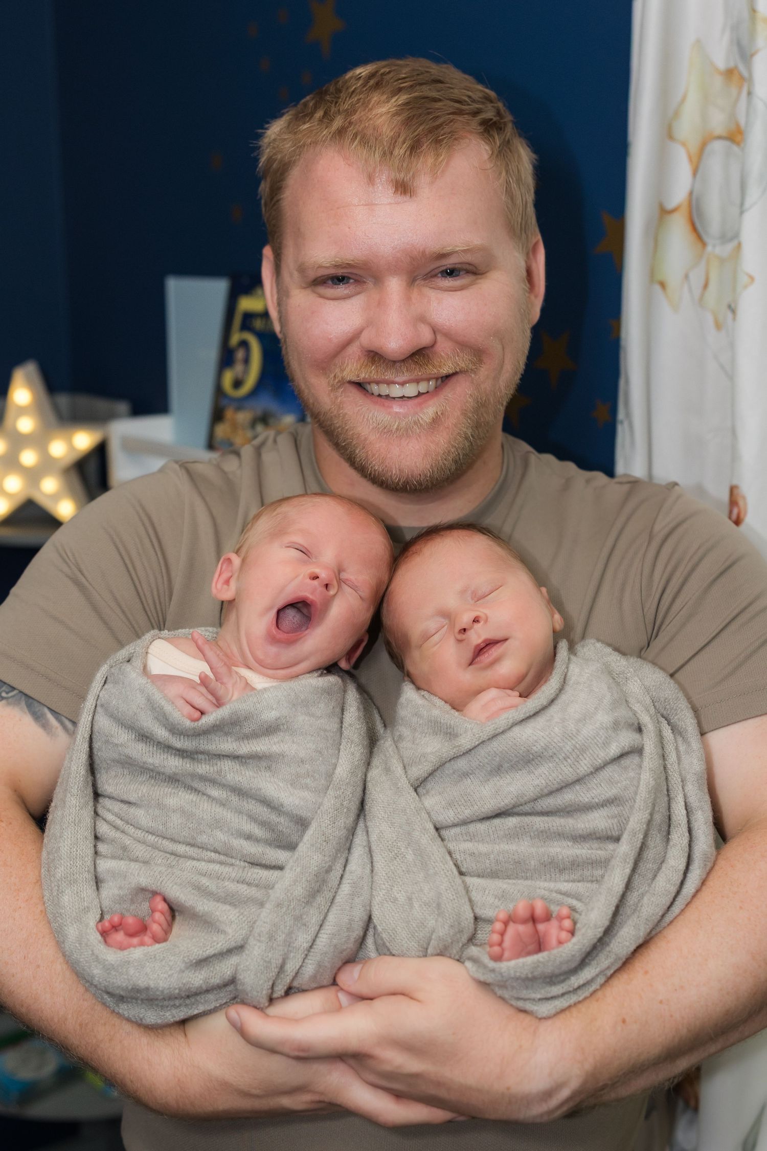 Smiling father holding two swaddled newborn twins, one yawning, in a decorated nursery room.