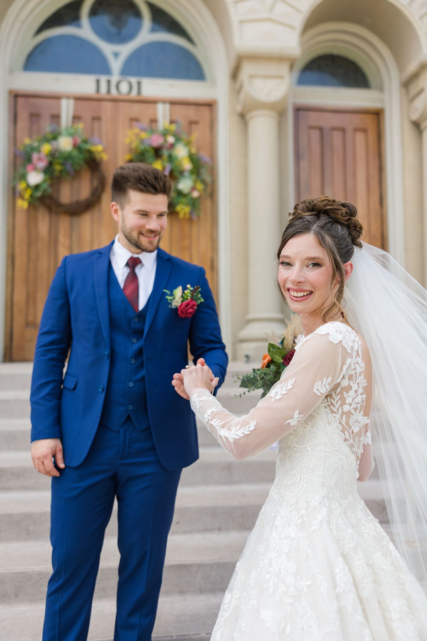 Smiling bride in lace gown holds grooms hand outside church doors wearing a blue three-piece suit.