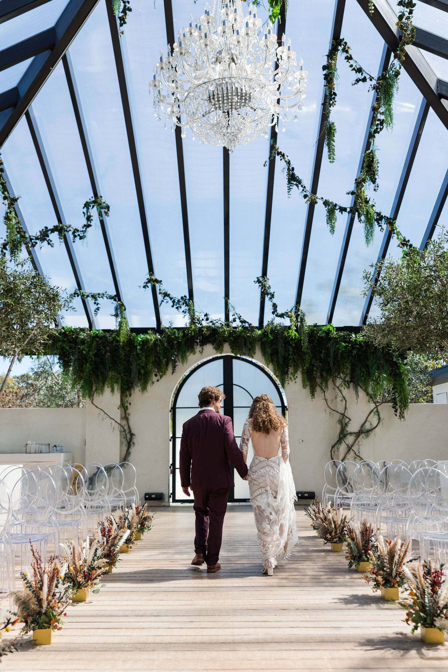 Bride and groom walk hand-in-hand down aisle in stunning glass greenhouse wedding venue with chandelier.