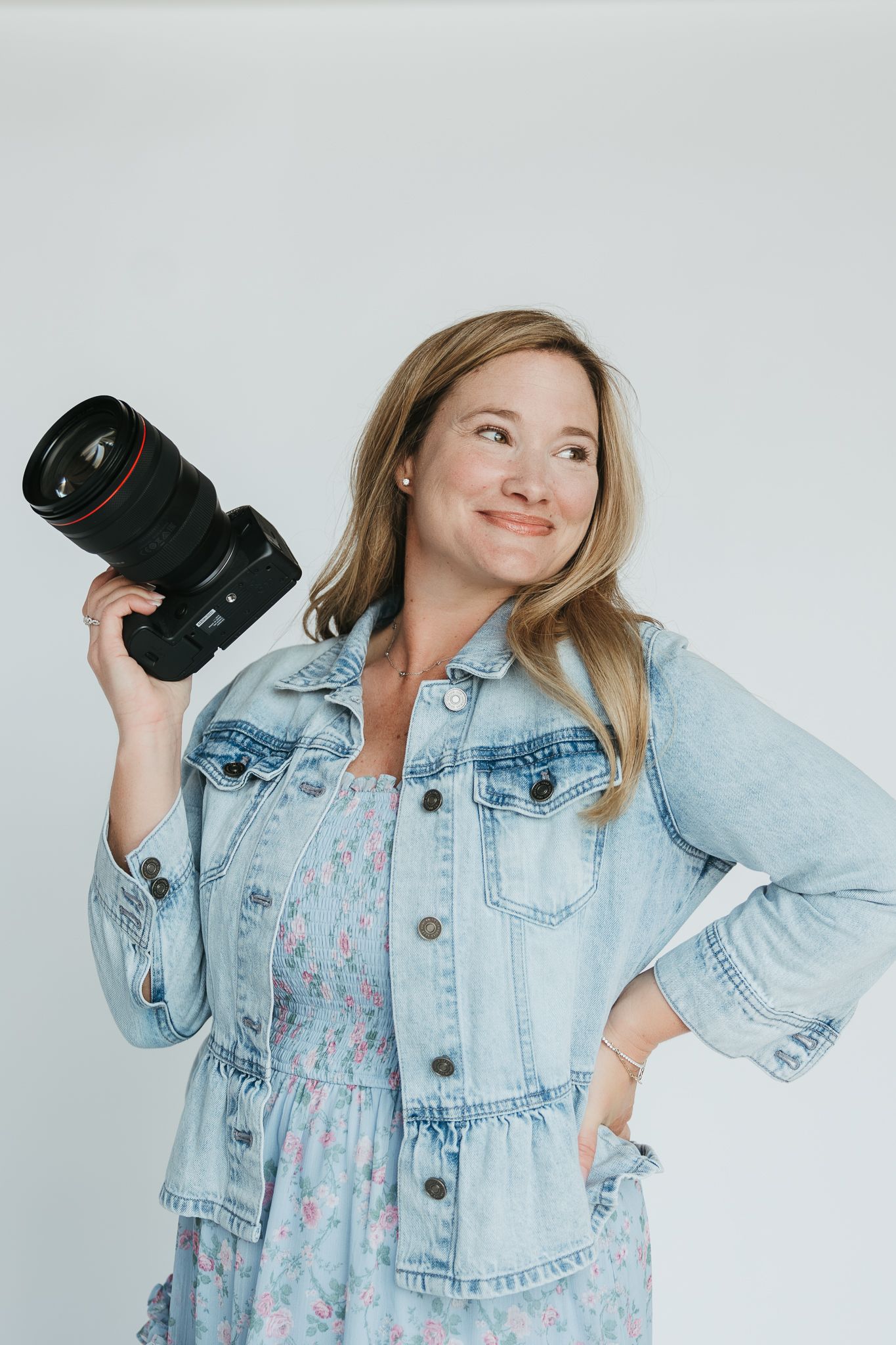 Photographer wearing light denim jacket shows camera lens with white studio background.