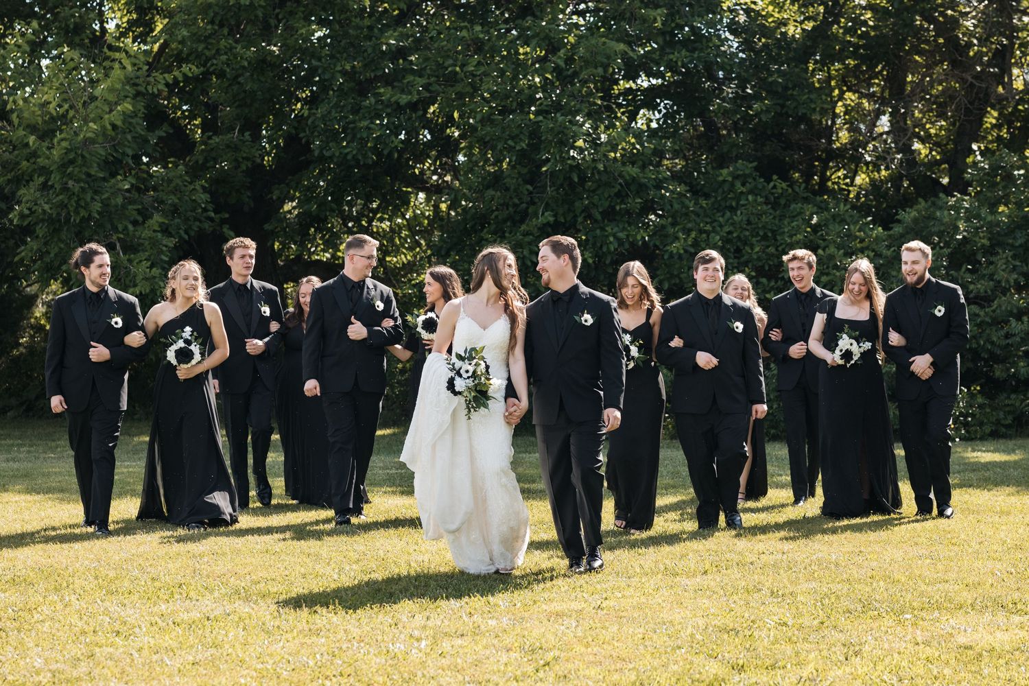 Wedding party walking together on grass with bride in white gown surrounded by attendants in black.