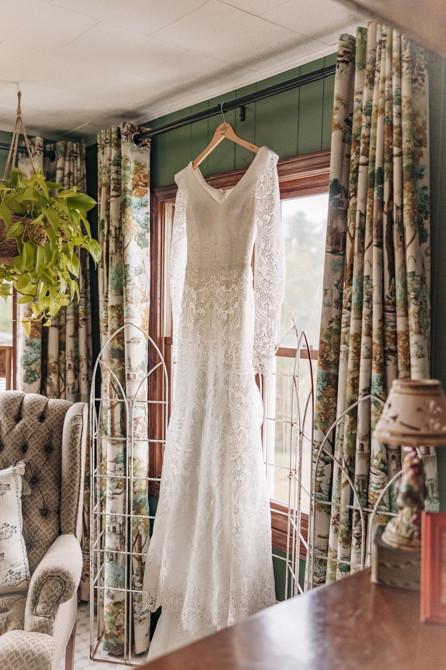 Elegant white lace wedding gown displayed in room with floral curtains.