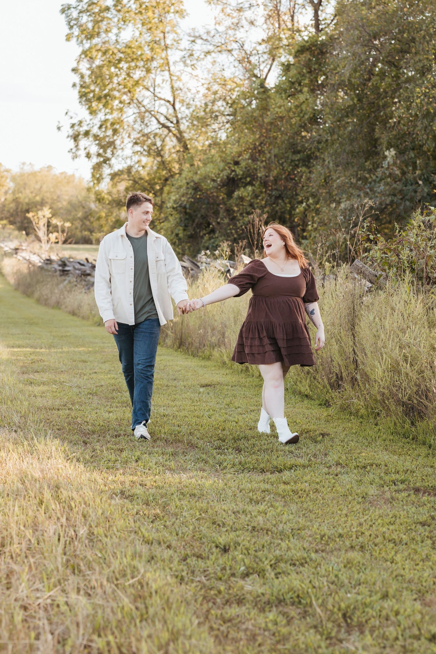 Couple holding hands while walking along a grassy path beside wild vegetation.