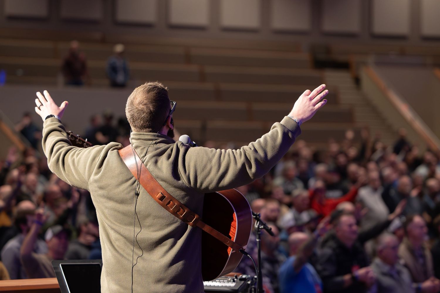 Performer in tan jacket stands with arms raised before large audience at concert venue.