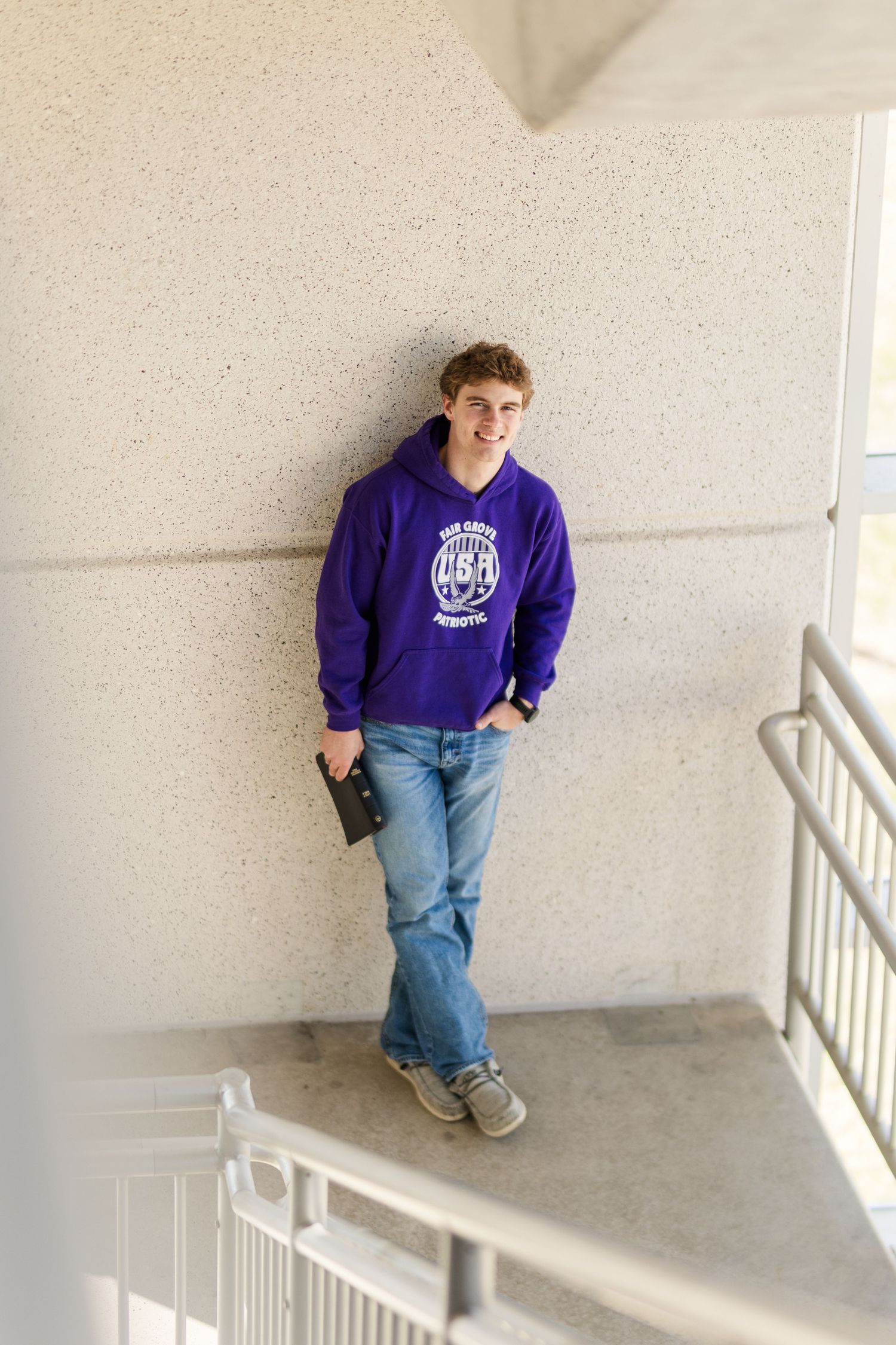 Person in purple sweatshirt and jeans leaning against white wall in stairwell.