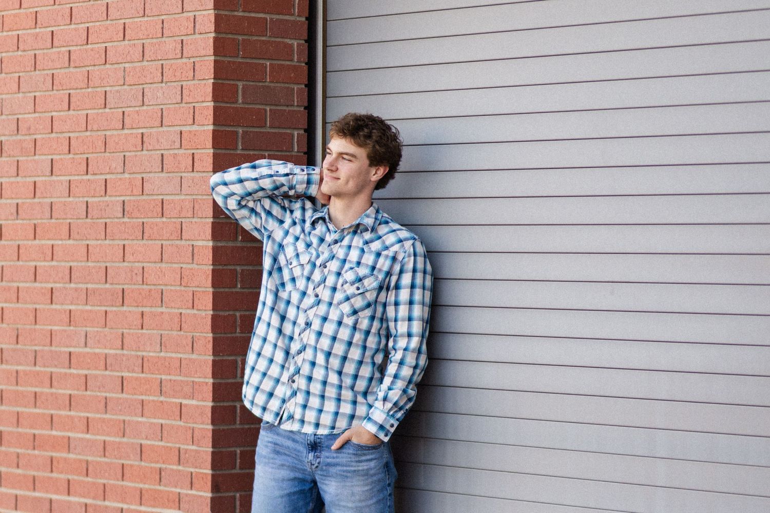 Someone in a blue plaid shirt and jeans leans against a brick wall with one hand behind their head.