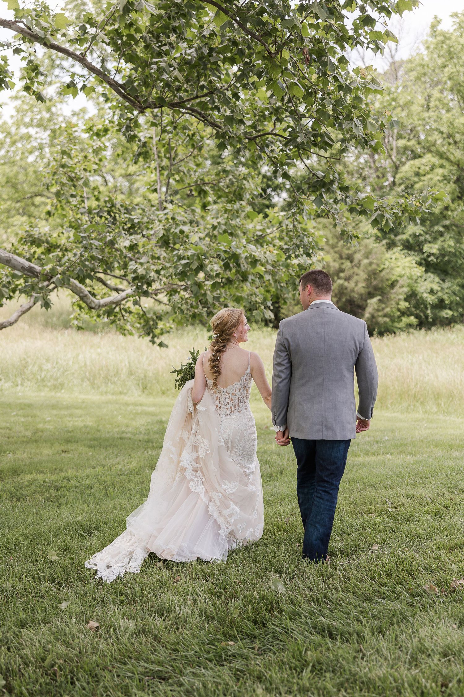 Couple in wedding attire walking together on lush green grass beneath trees on a sunny summer day.