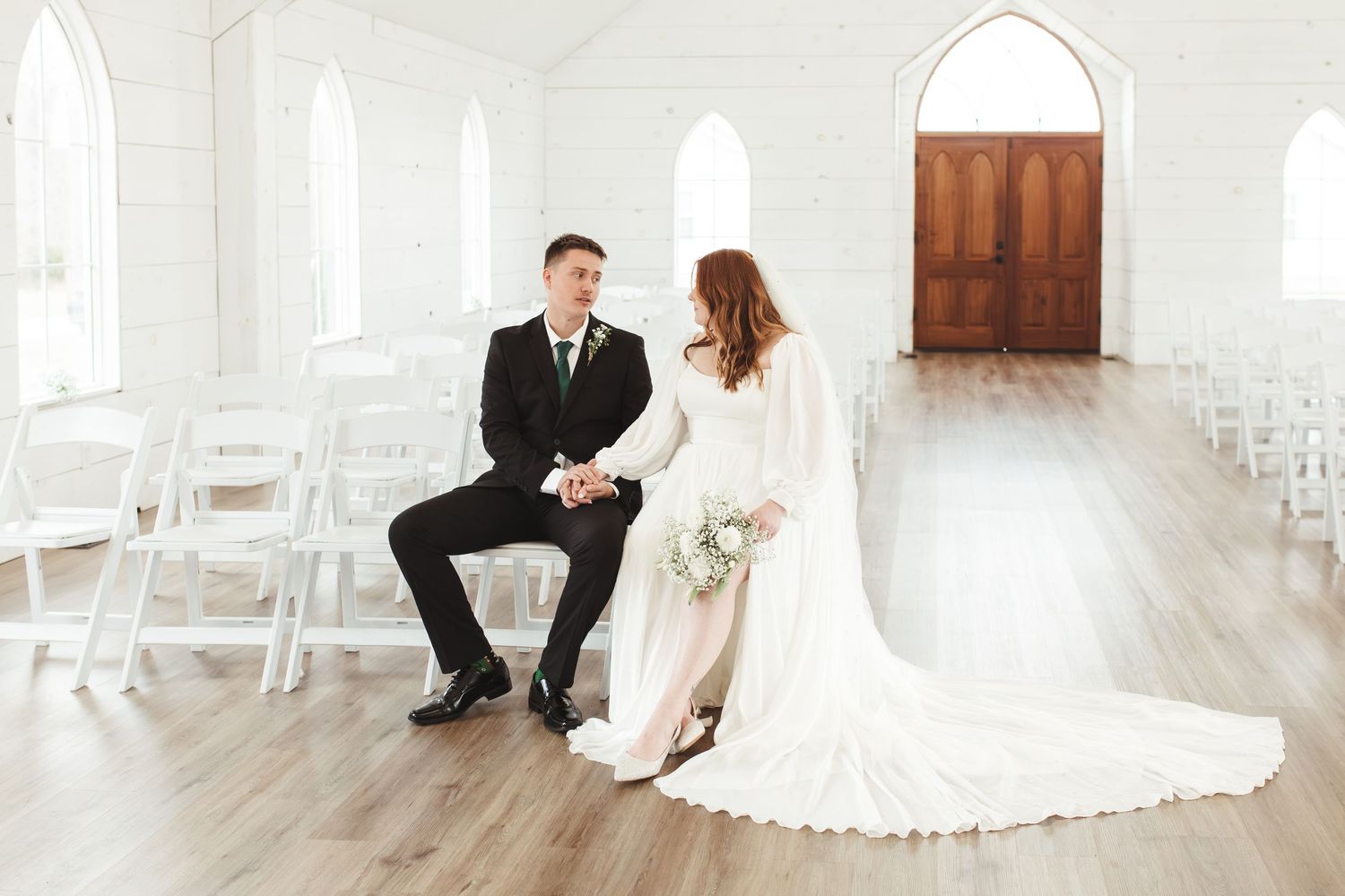 Bride and groom holding hands in a bright white wedding chapel with arched windows and wooden doors.